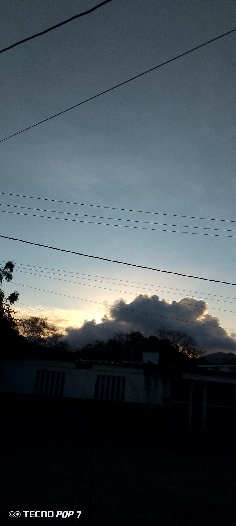 The image presents an outdoor landscape scene captured during either sunrise or sunset in Altagracia, Venezuela. The sky dominates the upper portion of the frame, displaying a gradient from a greyish-blue at the top to a lighter, more ethereal blue-grey towards the horizon. A bright, golden light source, presumably the sun, is visible behind a prominent, dark, and irregularly shaped cloud formation situated in the mid-ground. This cloud mass partially obscures the sun, creating a dramatic contrast.
Several thin, dark utility lines or power lines extend horizontally across the sky at different elevations, suggesting the presence of infrastructure. One thicker, darker line also runs diagonally from the upper left corner. Below the horizon, a continuous band of dark silhouettes reveals trees, dense foliage, and the roofs of buildings or houses. The structures in the foreground are largely in shadow, with visible window outlines featuring vertical bars. Faint outlines of distant mountains or hills can be observed in the far background, particularly towards the right side. The overall atmosphere is one of serene twilight or dawn. The text "TECNO POP 7" is visible in white in the bottom left corner of the image.