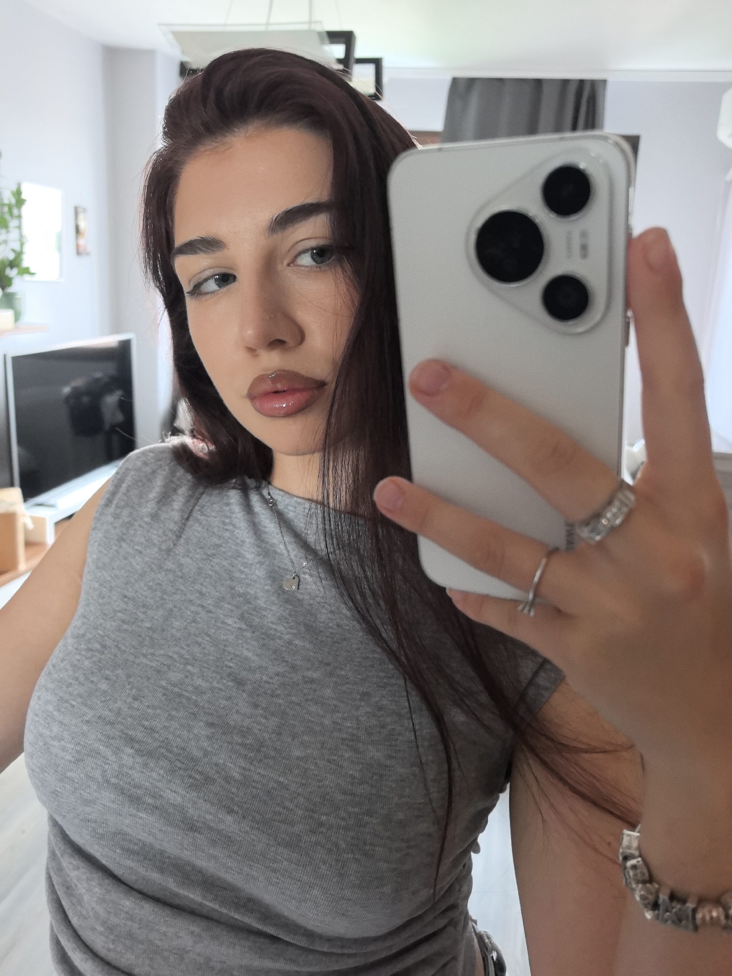 A young woman is depicted in a selfie, likely taken via a mirror reflection, in an indoor setting in Stara Zagora, Bulgaria. She has long, dark brown hair and a neutral to slightly pensive expression, with her gaze directed to her right. Her makeup includes glossy lips and defined eyeliner.
She is wearing a simple grey sleeveless top. A delicate silver necklace with a small heart-shaped pendant hangs around her neck. On her right hand, which holds the white smartphone, she wears several rings: two on her ring finger and one on her pinky. A silver-toned bracelet is visible on her left wrist.
The smartphone she holds is white with a prominent rear camera module featuring three circular black lenses arranged in a triangular pattern. The background shows a brightly lit interior, possibly a living room or bedroom, with a white wall, a dark television or monitor on a stand, and some light-colored furniture or shelving. A ceiling light fixture is also visible in the reflection. No legible text is visible on any object. The scene appears to be taking place during the daytime.