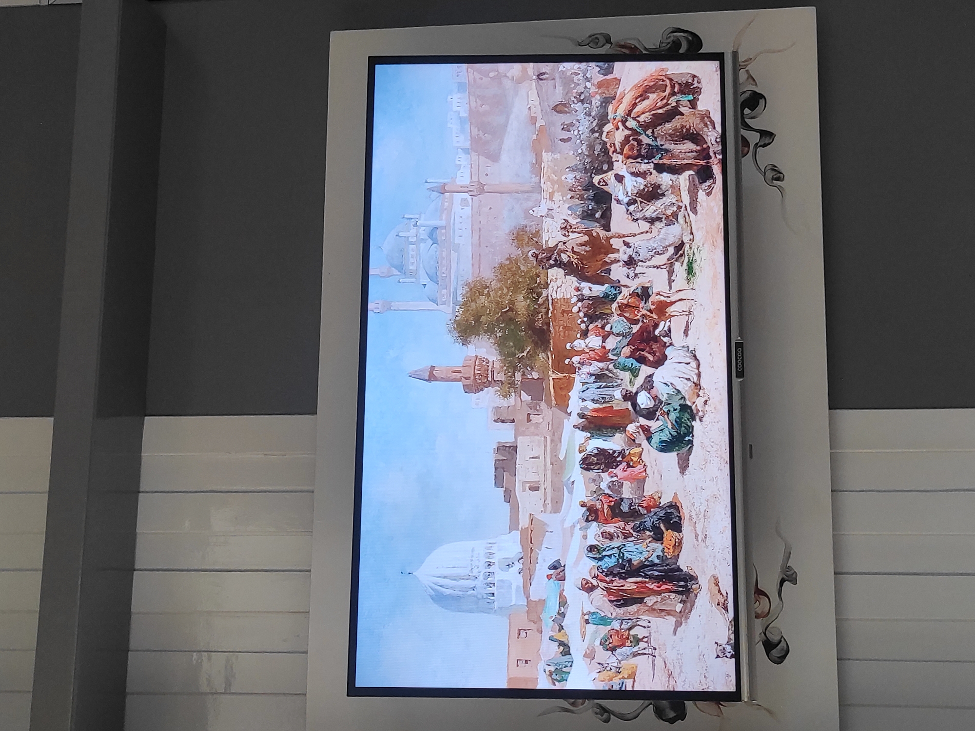 The image shows a modern flat-screen television displaying a painting. The painting depicts a bustling marketplace in what appears to be a Middle Eastern or North African city. In the foreground, numerous people in traditional attire are gathered, some standing, some sitting, and some tending to camels and donkeys laden with goods. The marketplace is filled with various stalls and tents, suggesting a vibrant commercial scene.
In the background, there are impressive architectural structures. A large white dome dominates the left side of the painting, likely part of a significant building such as a mosque or mausoleum. To the right of this, more buildings, including minarets, are visible, hinting at a city with a rich architectural heritage. The overall atmosphere of the painting is one of lively activity and historical significance.
The television itself is mounted on a wall that has a patterned design. The wall to the left of the television is covered in vertical white panels, while the wall behind and to the right of the television is a dark grey. The frame of the television is silver. Below the screen, there is a decorative border that features abstract, swirling black and white designs. On the bottom bezel of the TV, a small black logo with white text that reads "COOCAA" is visible.
The image does not contain any visual elements that would place it in the city of Sumedang, Indonesia. The depicted scene is clearly not of a location in Indonesia.
