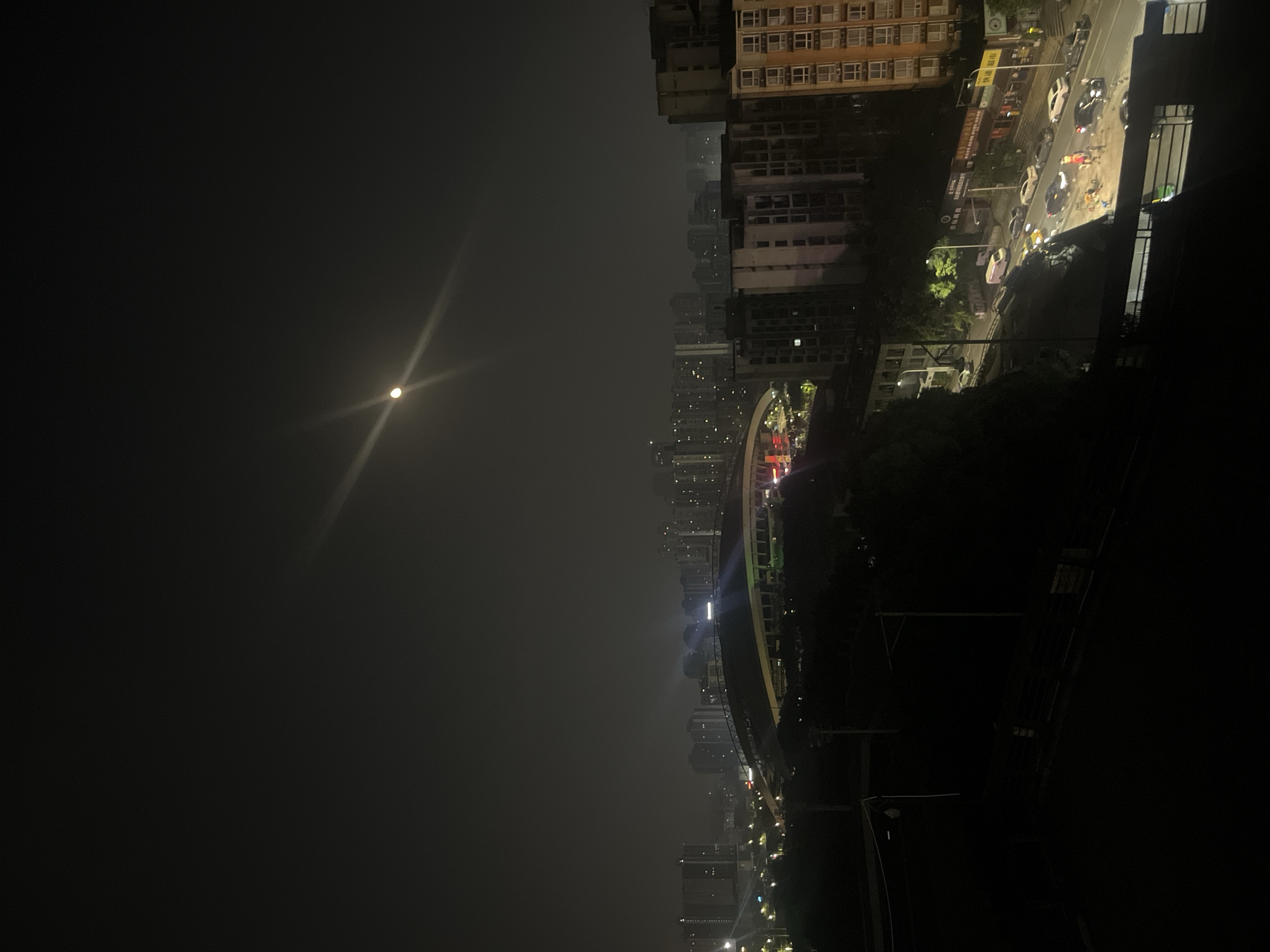 The image presents a nighttime panoramic view of an urban landscape in Jiulongpo District, Chongqing, China. The upper two-thirds of the frame consists of a dark, hazy sky featuring a bright, full moon exhibiting a pronounced starburst lens flare in the upper right quadrant.
Below the sky, a dense cityscape extends across the horizon, comprising numerous high-rise buildings, likely residential and commercial, with many illuminated windows, partially obscured by atmospheric conditions. In the mid-ground, a prominent architectural structure with a distinctive arched roof is visible, identified as the Chongqing Olympic Sports Center, illuminated with white and green lights. Further right in the mid-ground, additional multi-story buildings are present.
The foreground on the right is occupied by a multi-story apartment building with a brown and beige facade, displaying numerous lit windows. Below this building, a curved two-lane urban street is visible in the lower right quadrant. Along this street, multiple vehicles are present, including cars and a light-colored van. A cluster of activity is observed on the street, illuminated by several localized bright lights. Approximately five to seven individuals are discernible within this area, engaged in an unspecified activity, possibly related to roadside vending or a small informal gathering. Orange traffic cones are placed near this activity. Illuminated storefronts with Chinese characters, including "快速超市" (Convenience Store), line the street. Dark, dense foliage and an elevated embankment separate the street level from the elevated vantage point of the photograph.
