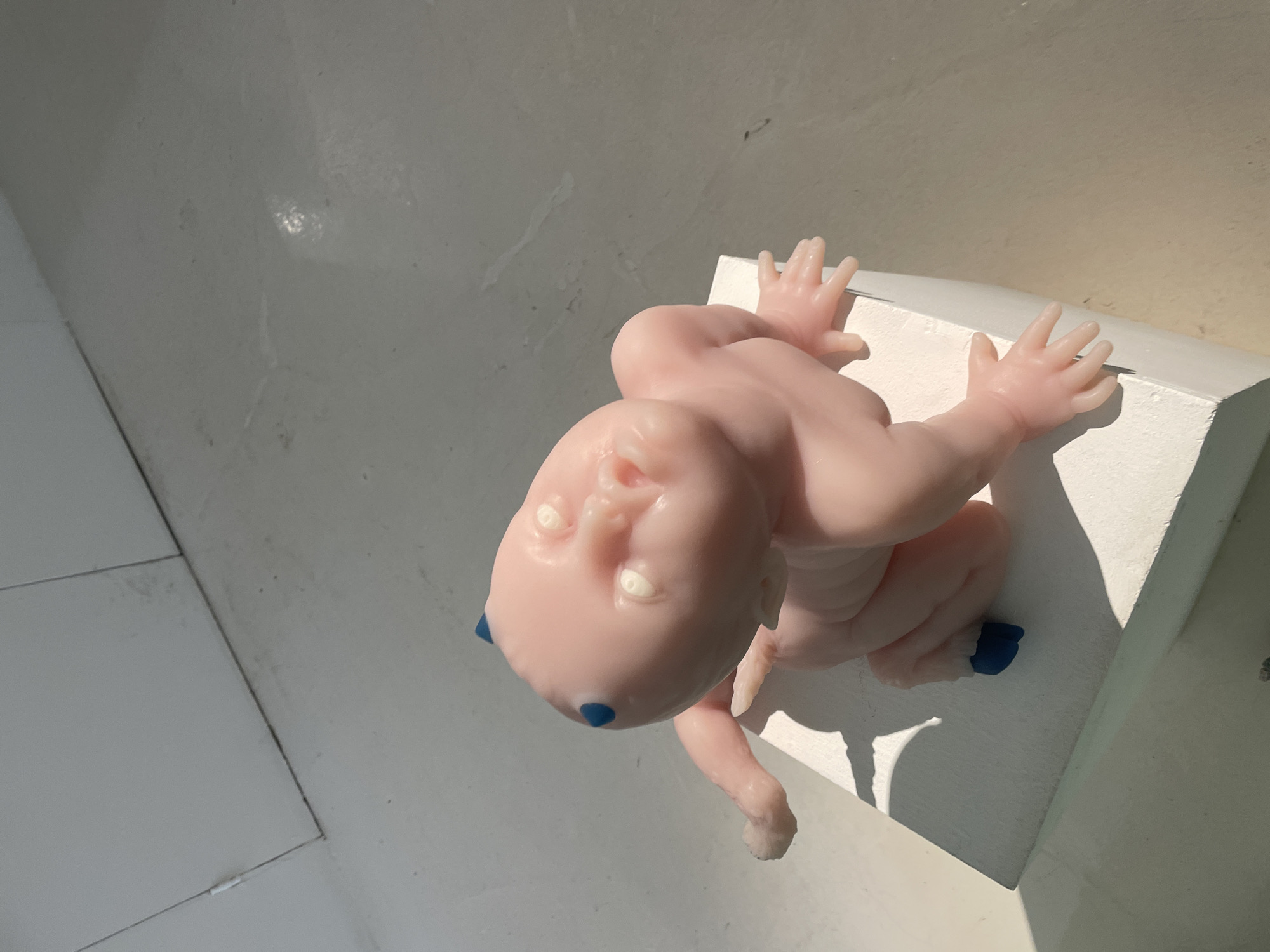 A close-up, overhead shot focuses on a pale pink, doll-like figure positioned on a white, rectangular surface. The figure, resembling a baby, is shown from the chest up, with its head tilted slightly to the left. It has a smooth, almost translucent skin tone, and its facial features are rendered with white eyes and a faint suggestion of a nose and mouth. Two small, round, dark blue marks are visible on the figure's forehead, just above its eyes. The figure's arms are bent at the elbows, and its hands are splayed with clearly defined fingers, resting on the white surface. The feet of the figure also appear to have dark blue accents. The lighting in the scene suggests it is daytime, with strong sunlight casting distinct shadows across the white surface and the figure itself, creating a high-contrast effect. The background is a pale, textured surface, possibly a wall or floor, with visible lines suggesting a tiled or paneled structure. The overall impression is of a still-life composition, with the doll as the central subject. No text is visible in the image.