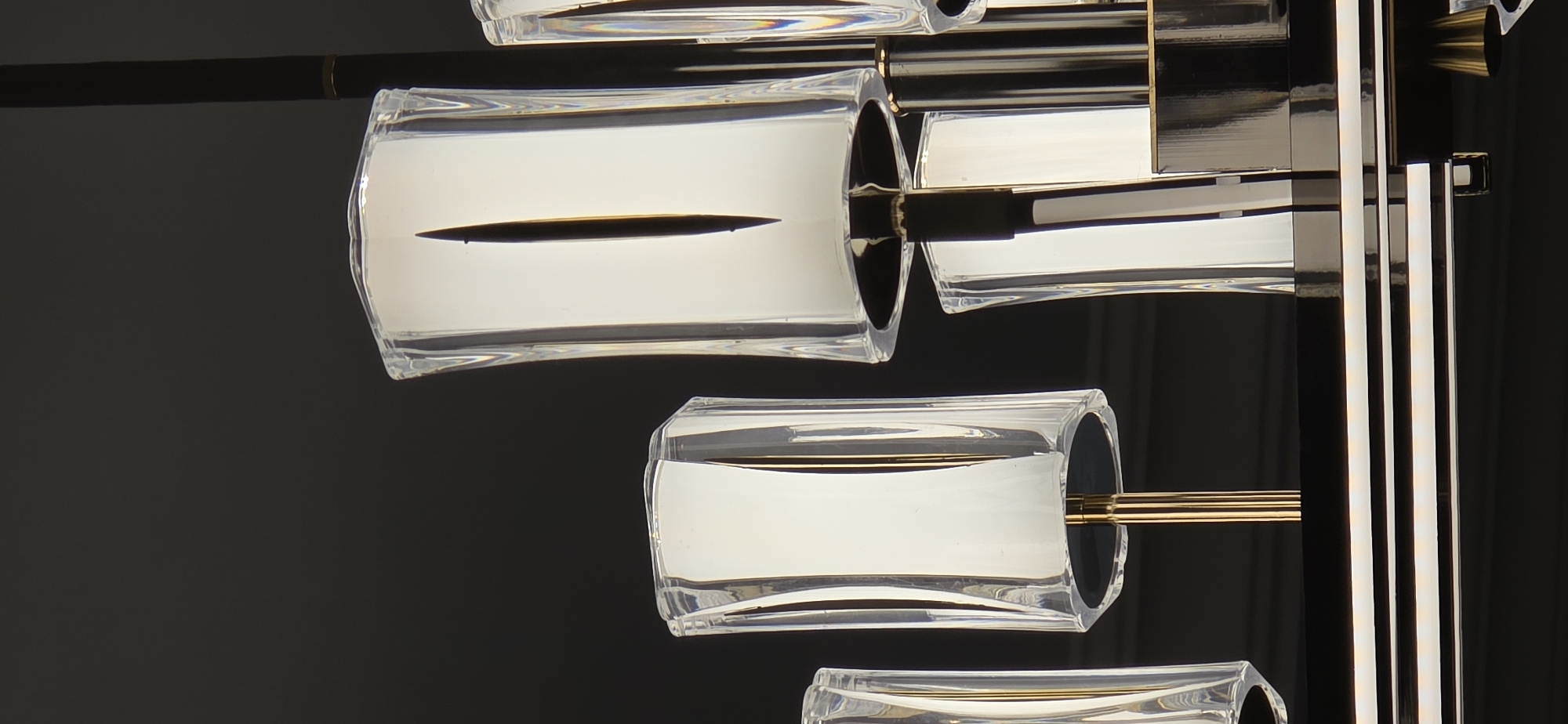 The image is a close-up, low-angle shot of a modern chandelier against a dark background. The chandelier features multiple glass shades, some appearing rectangular and others more cylindrical, with a frosted or opaque white interior that is illuminated from within. These shades are held by thin, metallic rods, possibly brass or gold-toned, which are attached to a larger, black structure that forms the base of the chandelier. The glass shades have a faceted or ribbed exterior, adding to their decorative appeal. Some of the shades have a distinct vertical element or shadow within them, suggesting a light source or internal design feature. The overall impression is one of contemporary elegance and sophistication. The image does not provide any information about a specific location in Sumgayit, Azerbaijan, as it is solely focused on the chandelier itself.