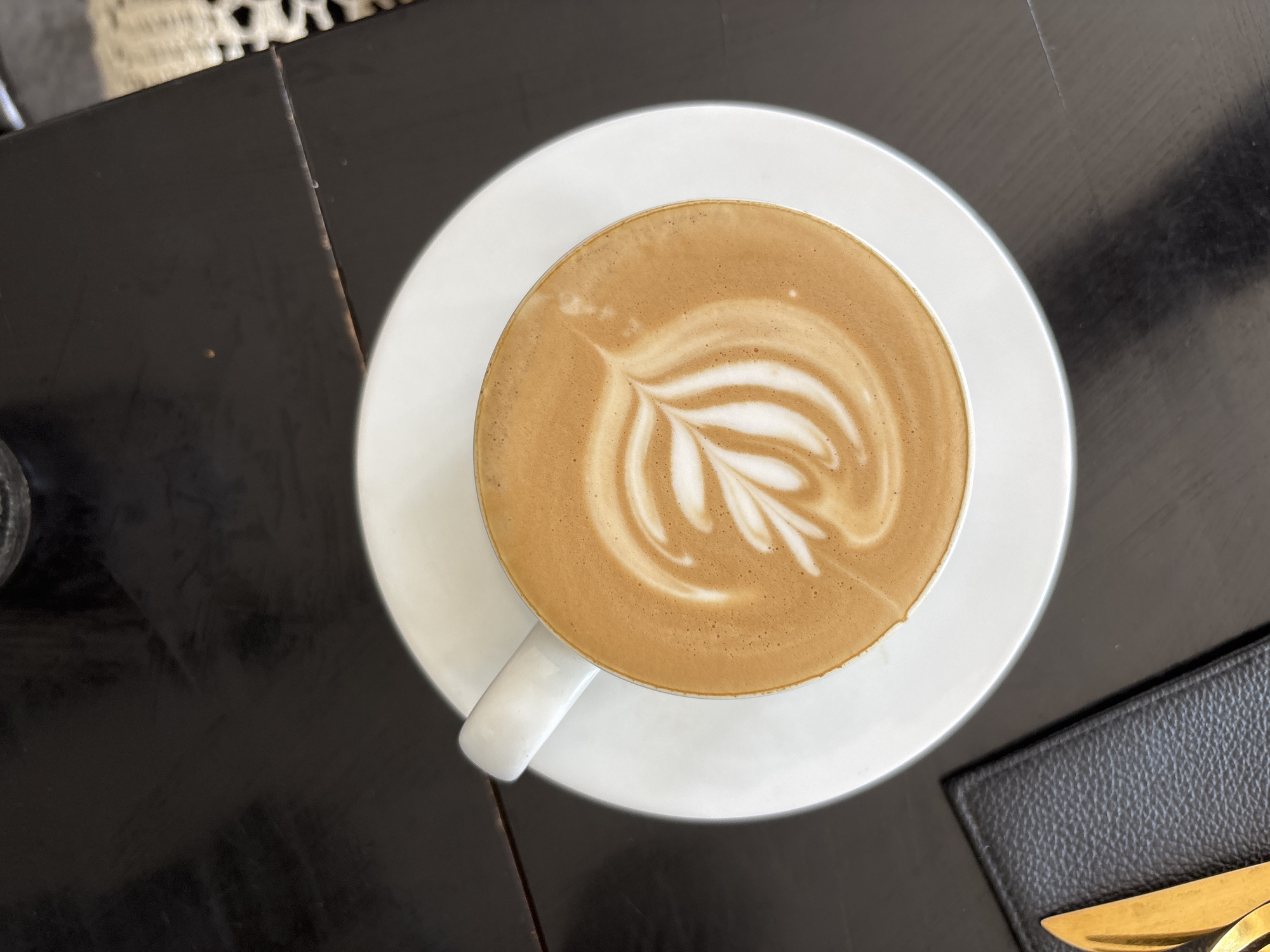 An overhead photograph captures a pristine white ceramic coffee cup, brimming with a latte or cappuccino, set upon a matching white saucer. The coffee's surface is adorned with delicate latte art, depicting a stylized white leaf or fern pattern against the rich brown of the beverage. The cup is centrally positioned on a dark, glossy wooden table, which exhibits subtle grain textures and minor signs of wear, including a visible seam towards the upper left.
In the top-left background, a soft-focused, light-colored object with a textured or woven pattern is partially visible, possibly part of a decorative rug or furniture. To the bottom right, a corner of a black, textured placemat or coaster is seen, upon which the golden-hued tips of two eating utensils – a knife and a fork – are resting, hinting at a dining context.
The scene conveys a calm and inviting atmosphere, focusing on the aesthetic presentation of the coffee. The lighting appears even, suggesting an indoor setting, likely a cafe or restaurant in Kuta Selatan, Indonesia. No individuals are present, and there is no visible text or direct indicators of the time of day or weather.