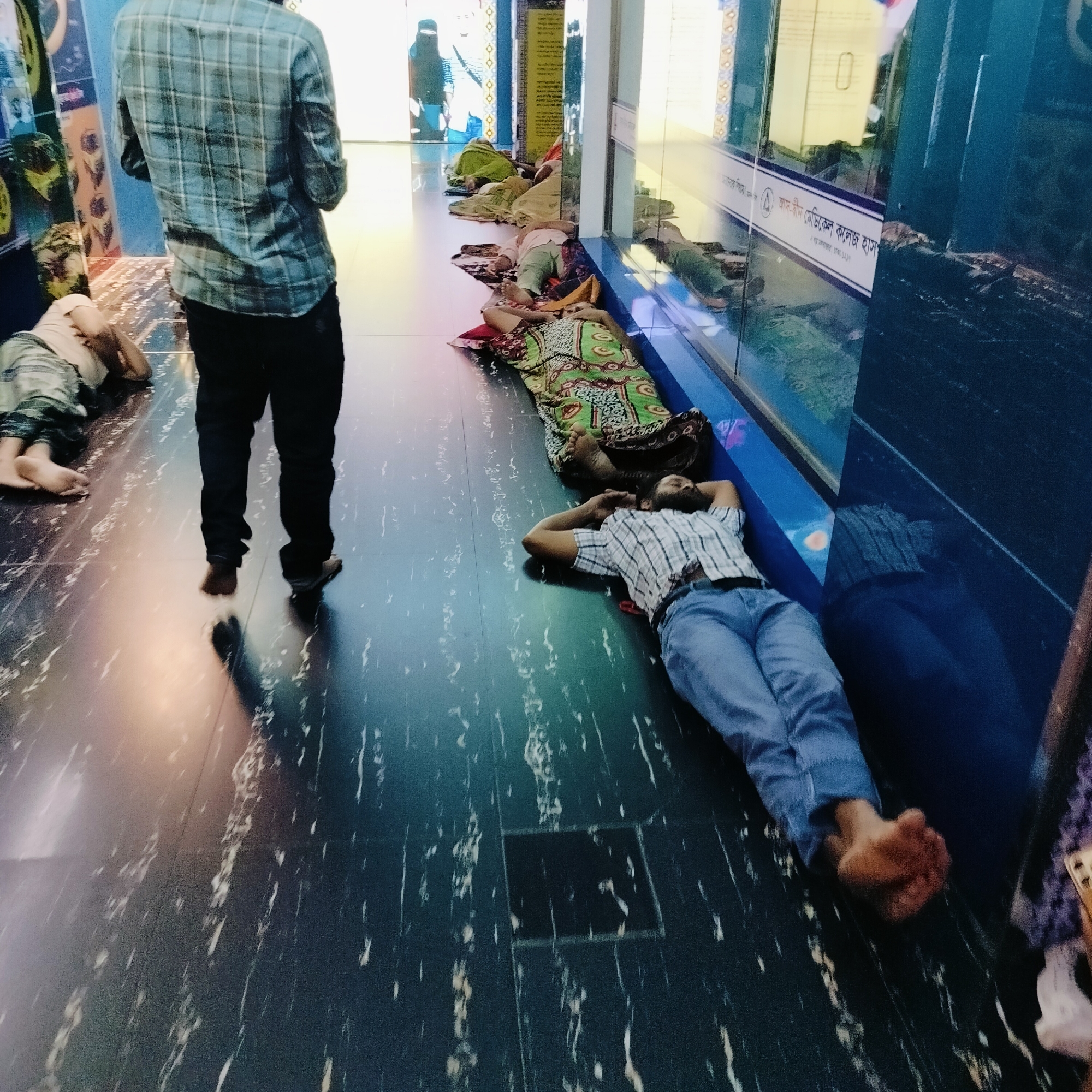 The image shows a scene within what appears to be a hospital or public building in Dhaka, Bangladesh. Several people are lying on the floor, some using coverings or pillows. One man stands in the foreground facing forward. The building has a shiny, reflective floor and a display window with signage written in Bengali. A person in a burqa is visible in the background through a doorway.