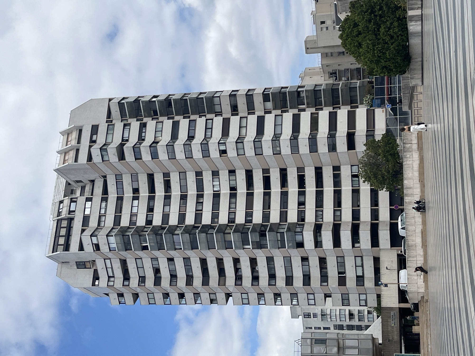 The image shows a tall, modern apartment building with a distinctive stepped facade in Créteil, France. The building is constructed with white and beige panels, and features numerous windows and balconies. The sky is partially cloudy. In the foreground, a paved public space with a striped pattern is visible. Several people are present, including a woman in white pants walking away from the camera, a man sitting on a low wall, and two individuals in a wheelchair. The background includes other buildings and trees. The scene appears to be a public square or pedestrian area adjacent to the apartment building.