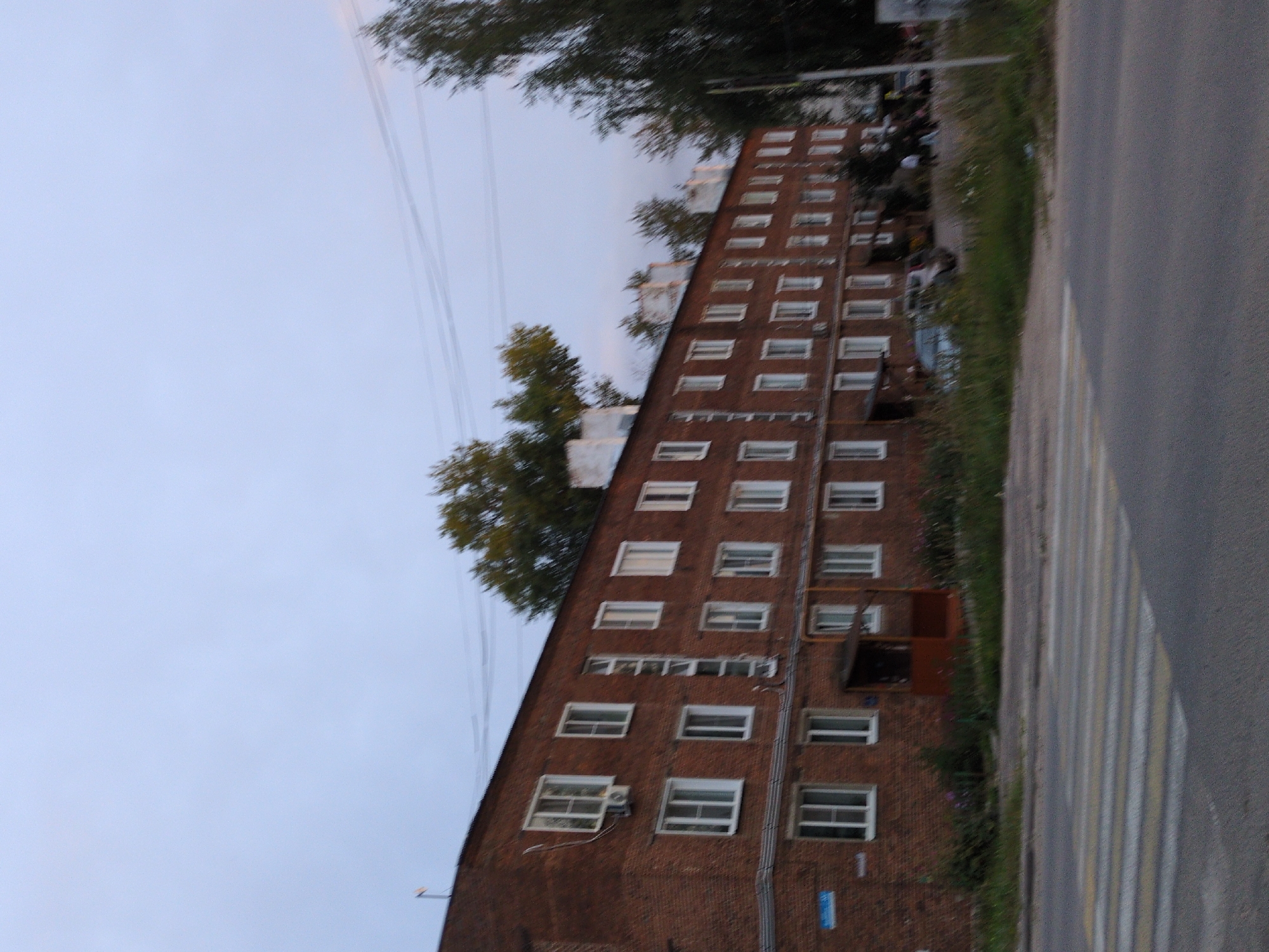 A wide shot captures a brick apartment building in Kulebaki, Russia, with a pedestrian crossing in the foreground. The building, painted a reddish-brown, features multiple rows of white-framed windows. An air conditioning unit is visible on the left side of the building. Trees, some with green foliage and others appearing sparser, are situated behind and to the right of the building. The sky above is overcast and pale blue. In the background, a few cars are parked and a couple of indistinct figures are visible. The road in front of the building has a striped pedestrian crossing marked with yellow and white paint.