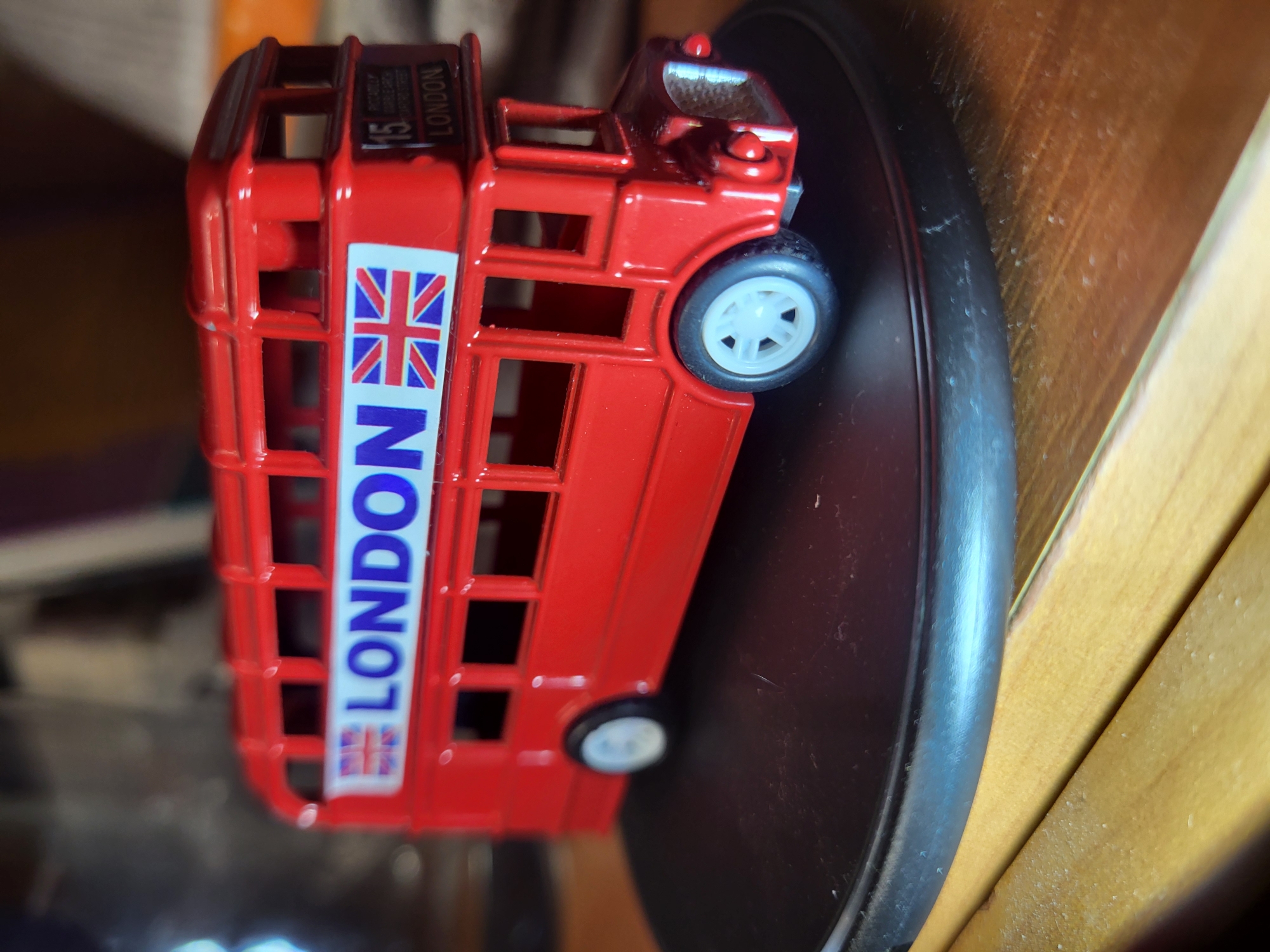 A red, double-decker toy bus is displayed on a black, circular base. The bus has the word "LONDON" printed in blue letters across its side, accompanied by a Union Jack flag. The background is blurred, showing a wooden surface and indistinct objects. The toy bus is positioned at an angle, with its front to the right of the frame. The scene is indoors, likely on a table. There are no people or animals present. The location of Horishni Plavni, Ukraine is not identifiable from the image.