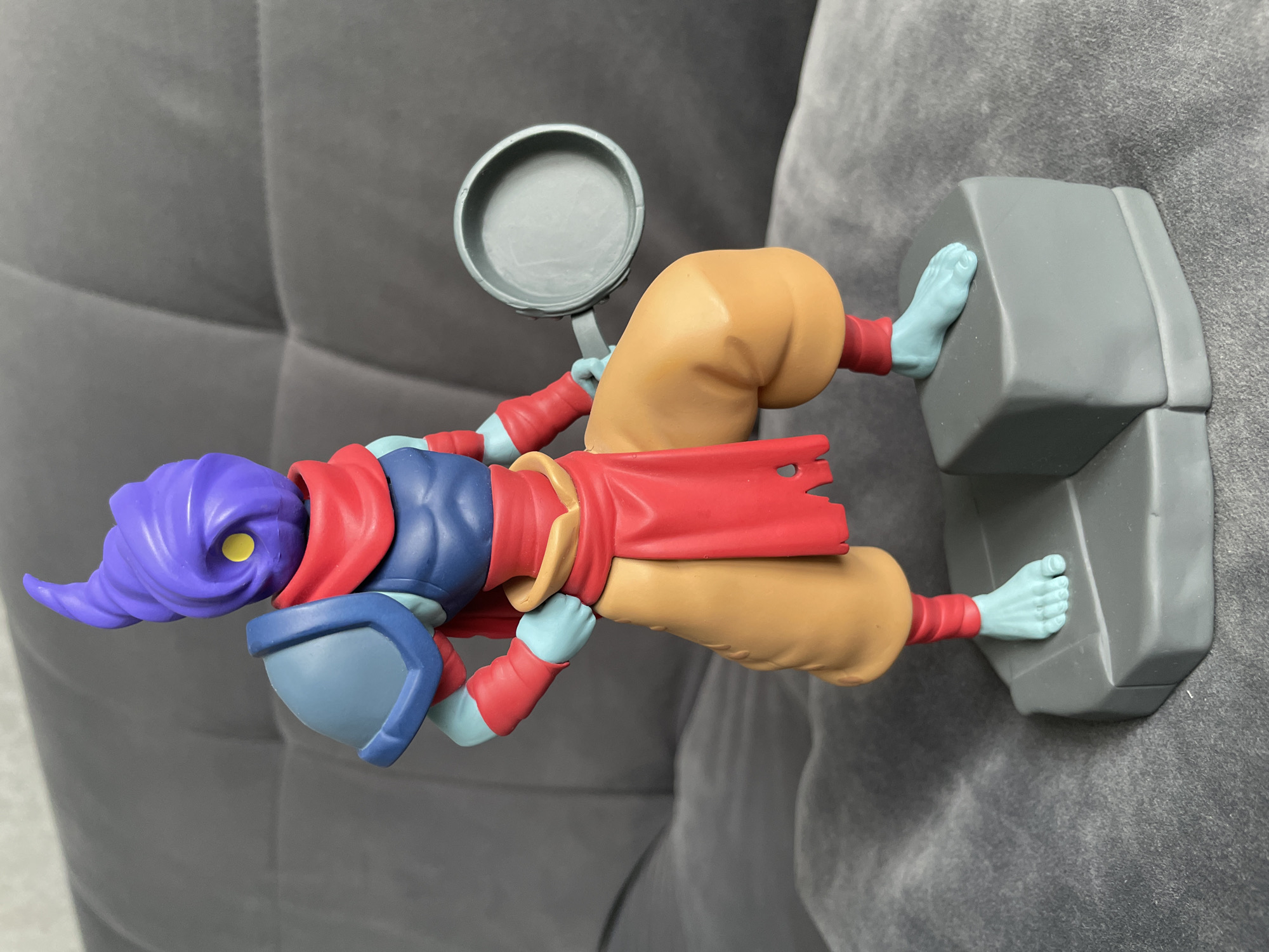 A figurine of a video game character stands on a grey platform. The figurine has a purple, coiled head with a yellow eye. It wears a blue chest piece, red armbands, and a red scarf. Its pants are tan and baggy, and its feet are light blue. The character is holding a grey frying pan in its right hand. The background is a grey, tufted surface, possibly a couch. The scene is not located in Moscow, Russia.