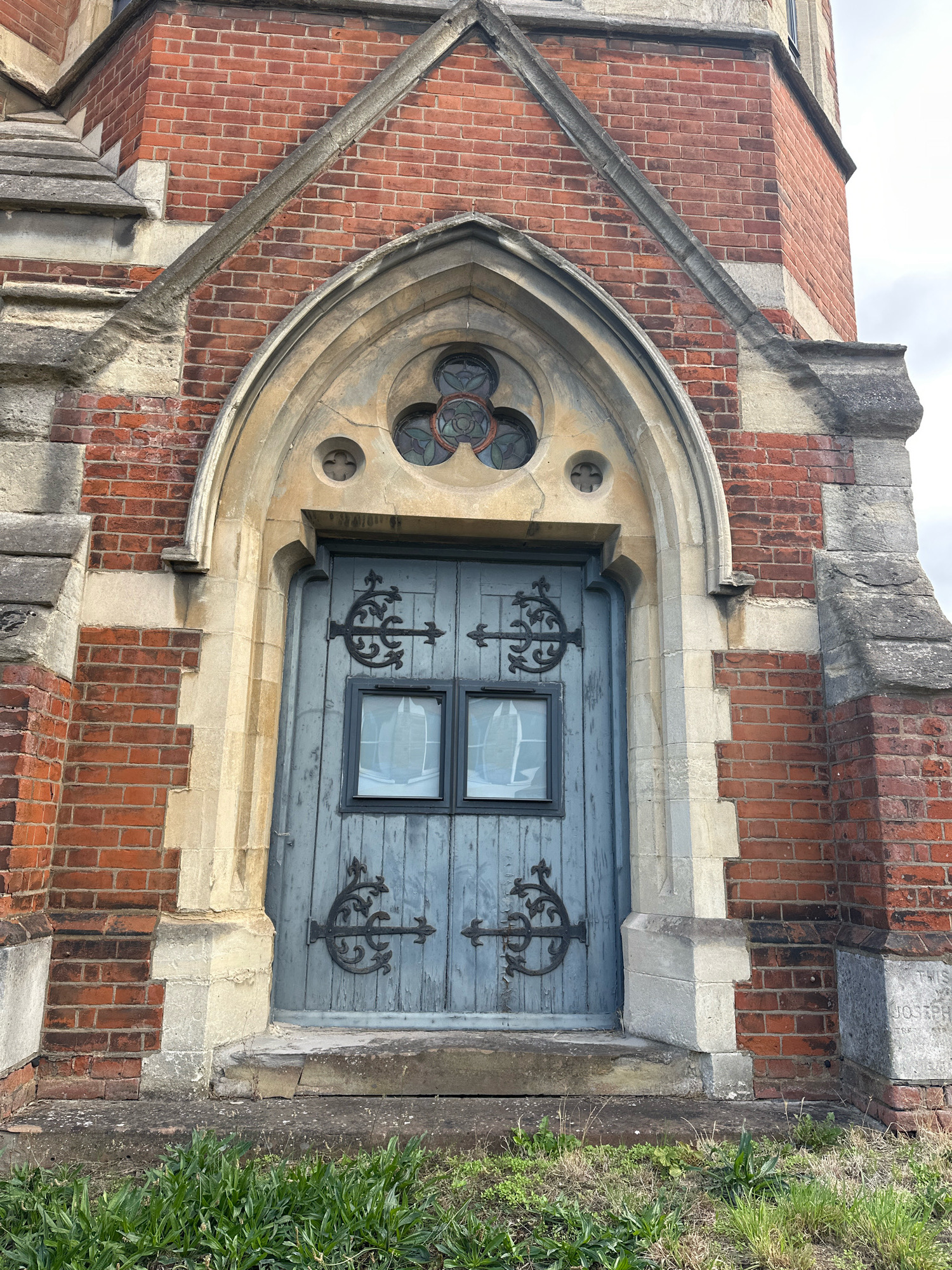 The image shows a close-up, eye-level, frontal view of a light blue, weathered wooden double door set within a gothic-style stone archway. The door has two square windows and ornate black metal hinges and latches. Above the door, within the arch, are three circular stained-glass windows, arranged in a trefoil pattern. The building's facade is constructed of red brick with contrasting grey stone detailing, particularly around the arch. The ground in front of the door is paved with rough concrete slabs, and overgrown green grass and weeds are visible at the bottom of the frame. The sky above is overcast. A faint inscription is visible on a stone block to the right of the door, which reads "THIS JOSEPH". This structure appears to be an entrance to a building with historical or religious significance. Based on the architectural style and the inscription, this could be a tomb entrance or a chapel, potentially within a cemetery.