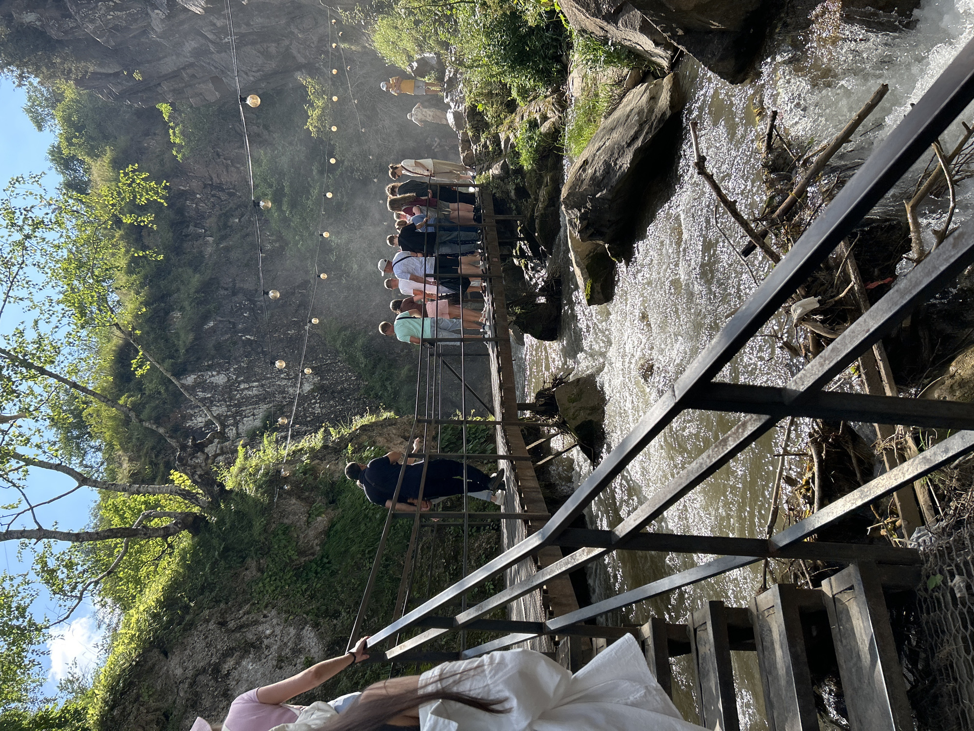 A group of people are standing on a metal bridge crossing a river. The bridge is made of wooden planks and has black metal railings. The river is flowing rapidly with white water, and there are rocks on either side. In the background, there is a rocky cliff face covered in greenery, and string lights are hung across the scene. The sky is blue and there are some trees visible. On the left side of the image, there are stairs leading up to the bridge, with more people on them. The overall scene appears to be a tourist attraction or a scenic spot. The location is identified as Krasny Kurgan, Russia.