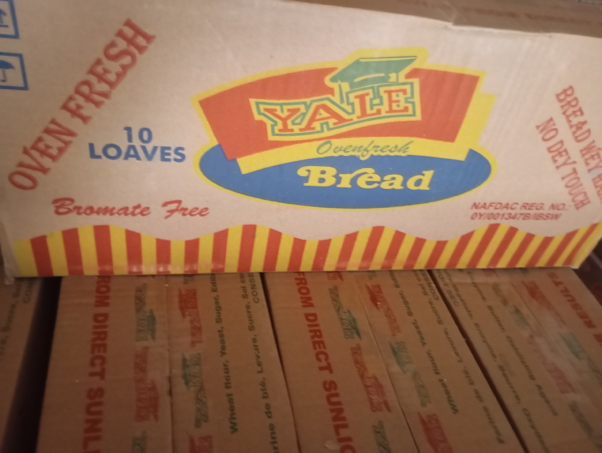 The image shows a stack of brown cardboard boxes containing "YALE Ovenfresh Bread" from Nigeria. The top box is labeled with "OVEN FRESH," "10 LOAVES," "Bromate Free," and the brand name "YALE" in red, yellow, and blue lettering. It also includes the tagline "Ovenfresh Bread" and the NAFDAC registration number. The sides of the boxes below show the text "FROM DIRECT SUNL" followed by ingredients in both English and French. The ingredients listed include Wheat flour, Yeast, Sugar. The scene appears to be a storage or distribution environment. The specific location within Wukari, Nigeria cannot be determined.