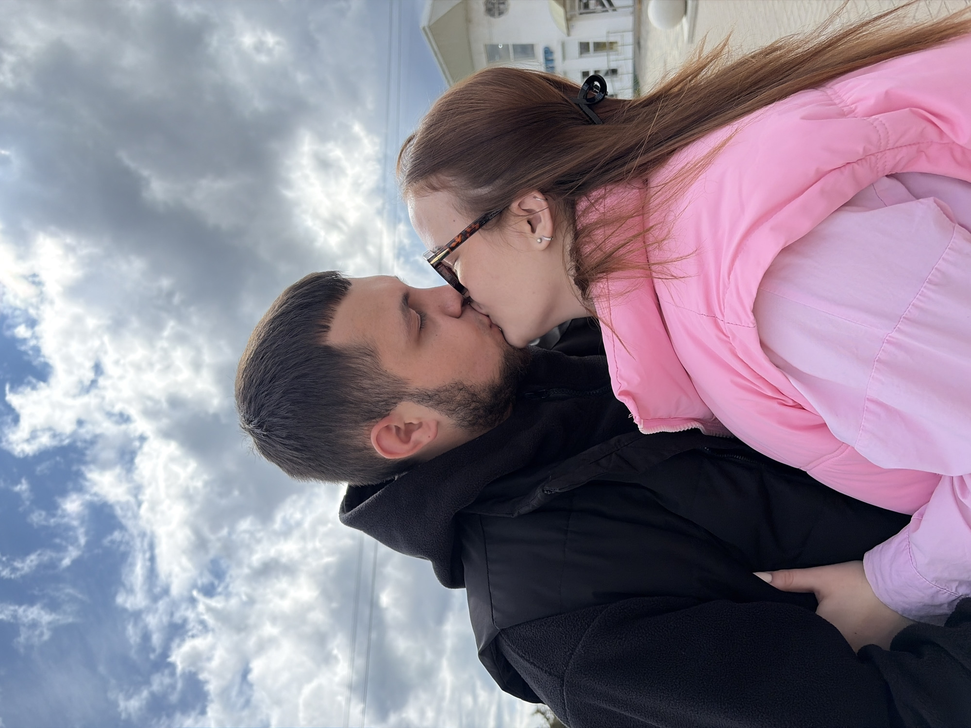 A man and a woman are kissing outdoors. The woman is wearing glasses and a pink jacket. The man is wearing a black jacket with a hood. The sky is overcast with clouds. The location appears to be Novopokrovskaya, Russia, with a building visible in the background.