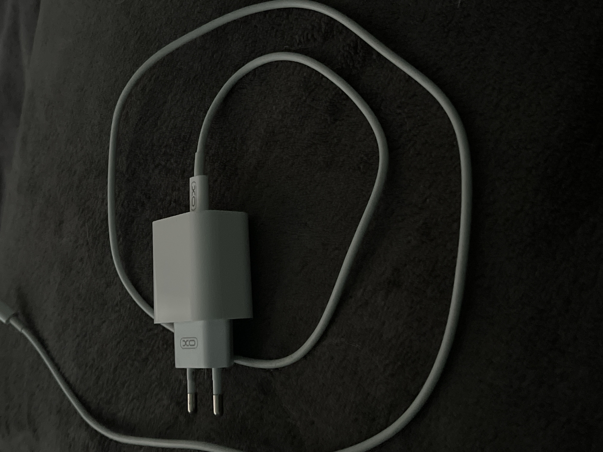 The image shows a white power adapter with two prongs and a white cable connected to it, resting on a dark, textured surface. The adapter has "XO" branding on one side. The cable is coiled loosely around the adapter. The scene is dimly lit, with the focus on the charger. There are no people or any indications of the specific location in Lviv, Ukraine.