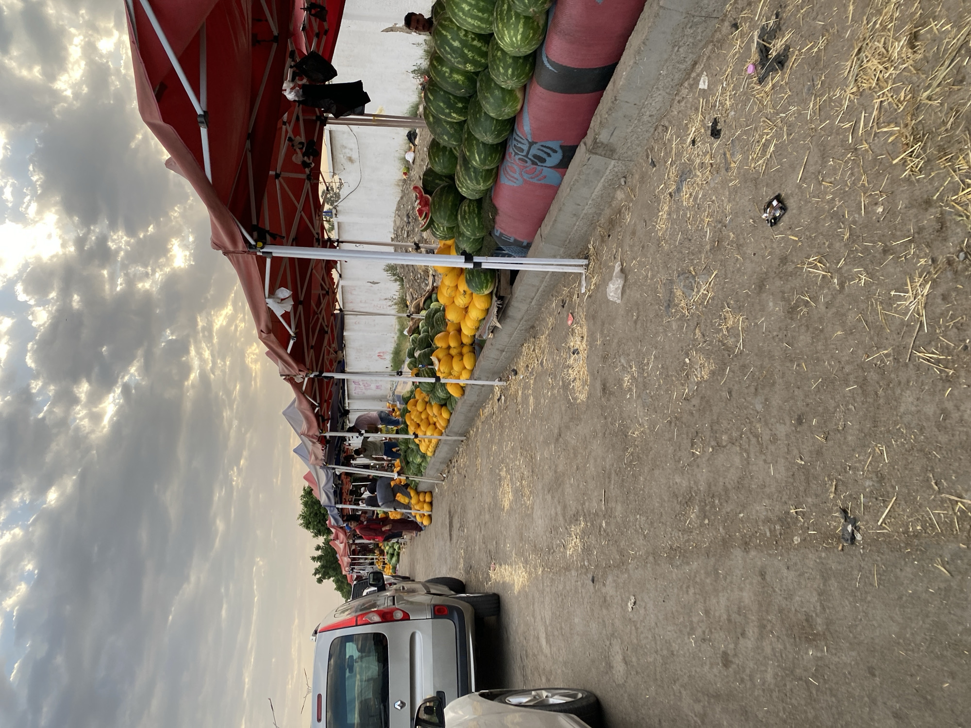 A street market scene is captured in Bougara, Algeria, under a cloudy sky. The market is lined with numerous stalls, covered by red and blue awnings, selling a large quantity of produce. Prominently displayed are rows of watermelons and yellow melons, arranged on wooden planks and concrete barriers. Several individuals are visible, some appearing to be vendors attending to the produce or interacting with unseen customers. A silver van is parked on the left side of the frame, and another white car is partially visible in the foreground. The ground is a dirt path, sparsely covered with dry straw and scattered debris, suggesting a typical outdoor market environment. The overall impression is of a busy, yet somewhat unkempt, marketplace.