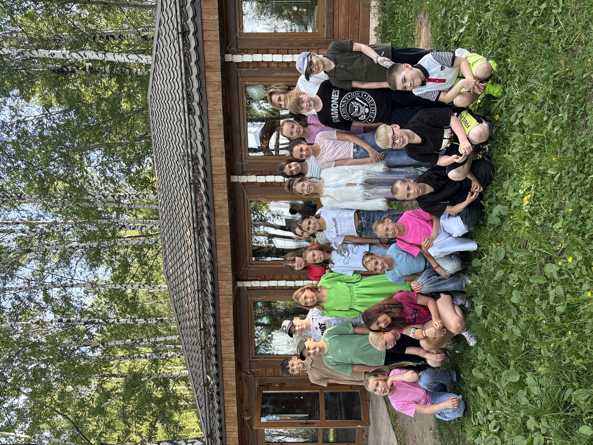A group of 21 people are sitting or kneeling on the grass in front of a one-story wooden building. The building has a brown roof and several windows. The setting appears to be outdoors, with birch trees visible in the background. It is difficult to confirm the location is specifically in Chaikovsky, Russia, without more contextual clues from the image itself.