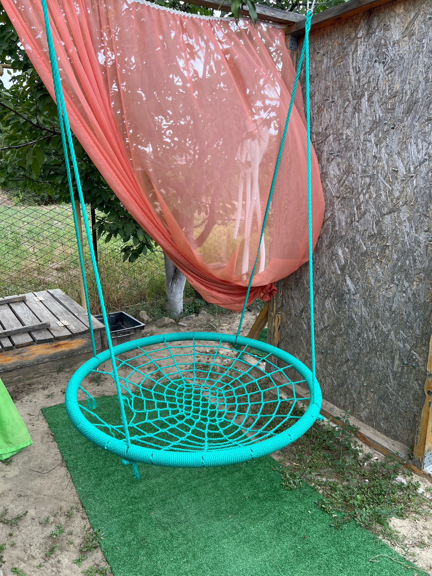 A turquoise spiderweb-style swing hangs from a wooden frame covered by a sheer coral-colored curtain. The swing is suspended by two turquoise ropes, attached to a horizontal beam. To the left of the swing, a weathered wooden structure with visible hinges sits on the ground. A black plastic container is visible behind the wooden structure. A patch of artificial grass lies beneath the swing. The background shows trees, a chain-link fence, and a wall constructed of oriented strand board. The scene is set outdoors.