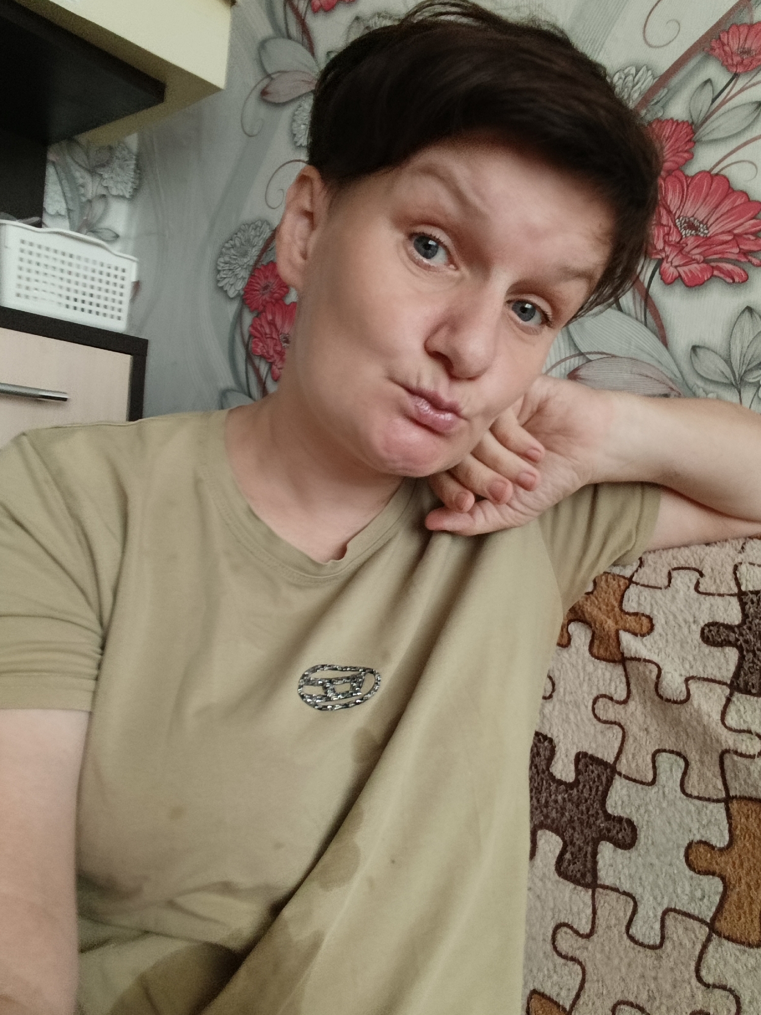 A woman with short brown hair and blue eyes is looking at the camera, resting her head on her hand. She is wearing a light brown t-shirt with a circular emblem on the left chest. The background shows a wall with floral wallpaper and a white basket on a shelf. The woman is sitting on a couch with a puzzle-piece patterned blanket.