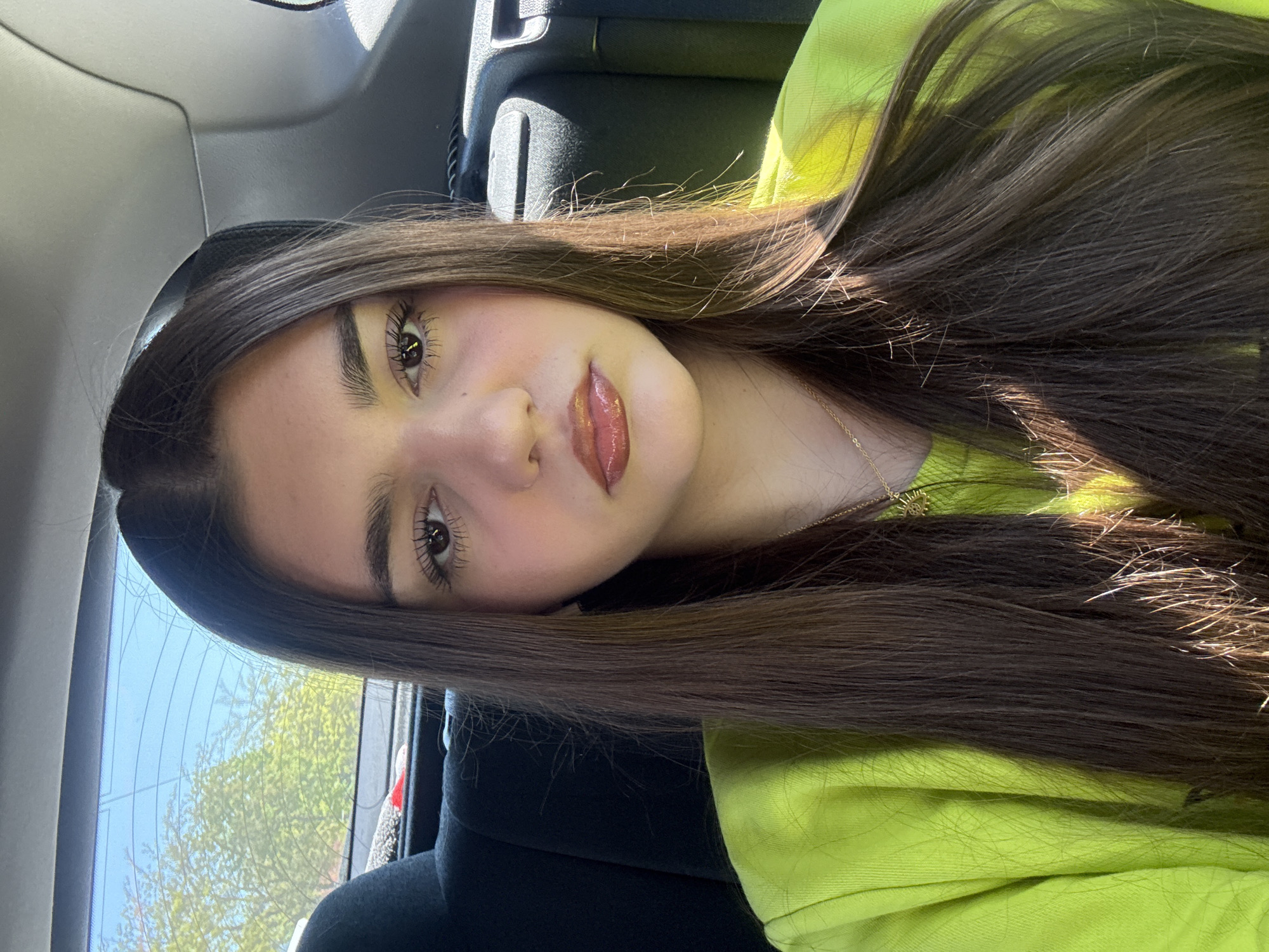 A young woman with long, brown hair, fair skin, and brown eyes is in the backseat of a car. She is wearing a bright yellow shirt and a gold necklace. The car's interior is black. Trees and a blue sky are visible through the car's window. Location: Unknown, Unknown.
