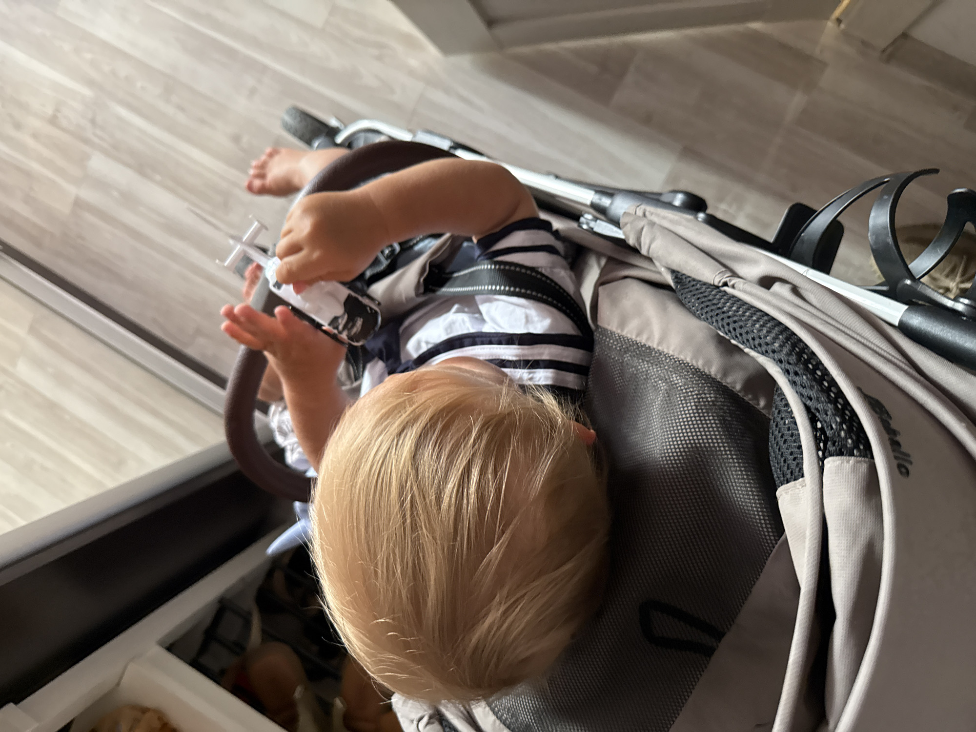 The image shows a toddler with blonde hair sitting in a stroller. The toddler is wearing a striped t-shirt and is holding a white toy that resembles a toy airplane or drone. The stroller is a neutral gray color and appears to be made of fabric and metal. The background shows a wooden floor, suggesting the scene is indoors. The lighting is natural, implying it is daytime. The image is taken from a high angle, looking down at the child. The overall mood seems relaxed and casual. The visible text on the stroller, though partially obscured, reads "Bugaboo". The specific location context is Sochi, Russia.