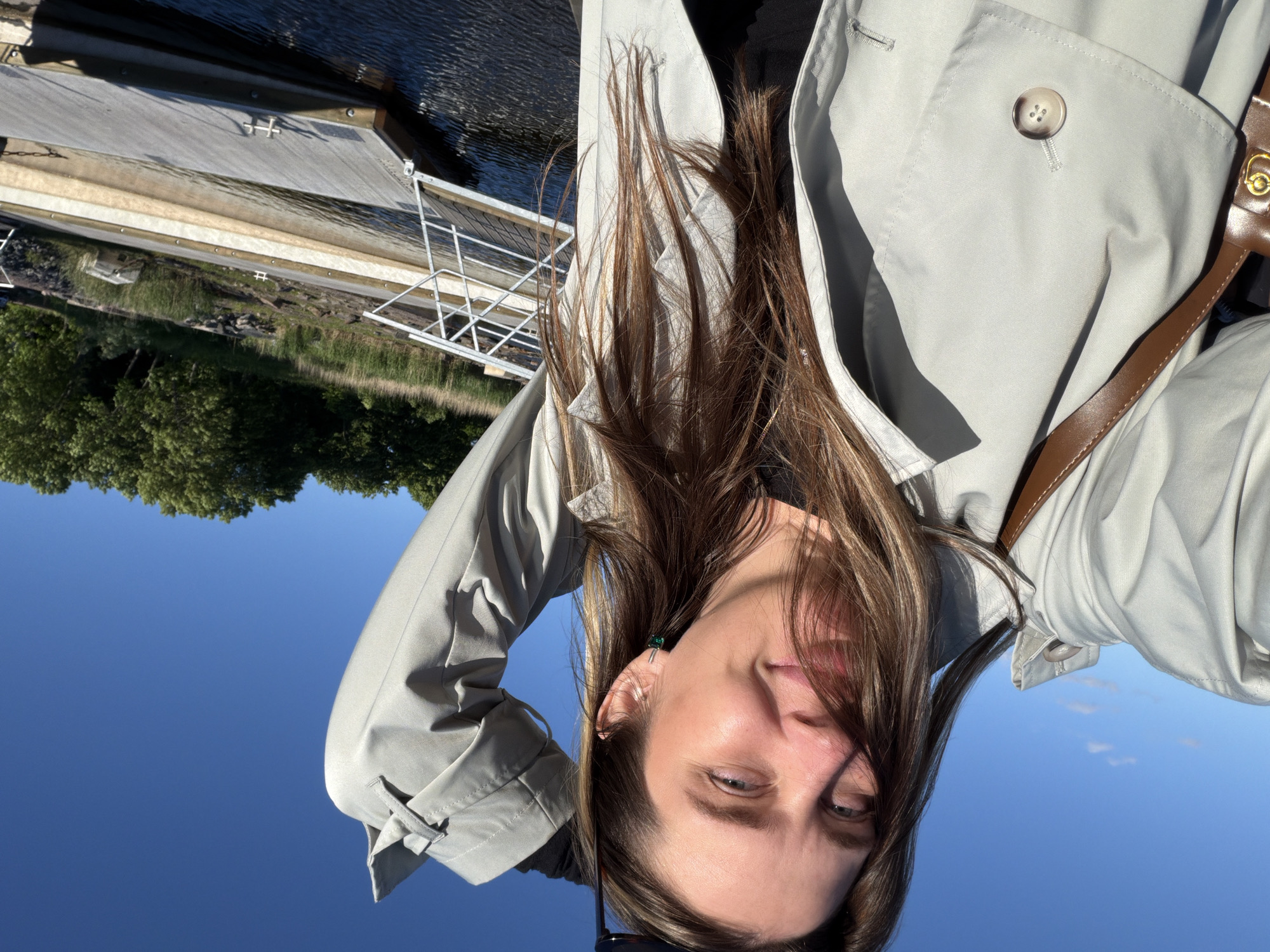 A woman is taking a selfie outdoors with a clear blue sky. She is wearing a light gray trench coat and a brown purse strap is visible over her shoulder. Her hair is long and brown. She is smiling and has her right arm raised, touching her hair. In the background, there is a body of water with docks and trees on the far bank. The image does not provide enough information to determine a specific location within Sortavala, Russia.