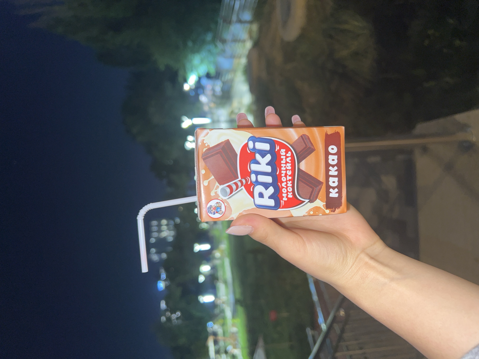 A person's hand is holding a small chocolate-flavored milk drink box with a straw inserted. The box has a cartoon image of a boy with a straw and illustrations of chocolate bars and milk. The text on the box is in Russian and says "Riki молочный коктейль какао". The background is a blurry outdoor scene at night, with lights from buildings and trees visible, suggesting an urban park or public space in Osh City, Kyrgyzstan. The lighting conditions indicate it is nighttime.