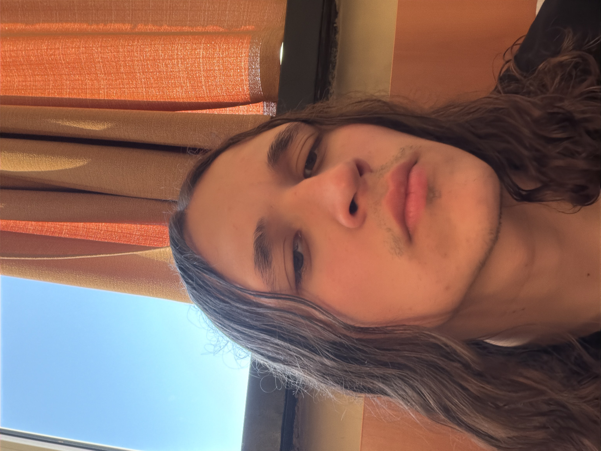 The image displays a close-up, sideways selfie of a young person with long, dark, wavy hair and light skin, captured in Plovdiv, Bulgaria. The person's face is prominent, showing one visible eye looking towards the camera, a neutral expression, and soft, full lips. Faint stubble or emerging facial hair is visible above the upper lip and on the chin. The original image orientation suggests the person is either lying down or tilting their head significantly.
The background reveals an indoor setting with elements suggesting proximity to a window. On what appears to be the left and upper side of the frame (due to the image's rotation), layered orange and tan curtains or blinds are visible, with sunlight highlighting their texture and folds. Below these curtains, a clear, vibrant blue sky is visible, indicating a sunny daytime scene with fair weather. A dark window frame partially separates the interior elements from the sky. No text is visible in the image. The overall impression is one of calm, perhaps taken during a moment of relaxation.