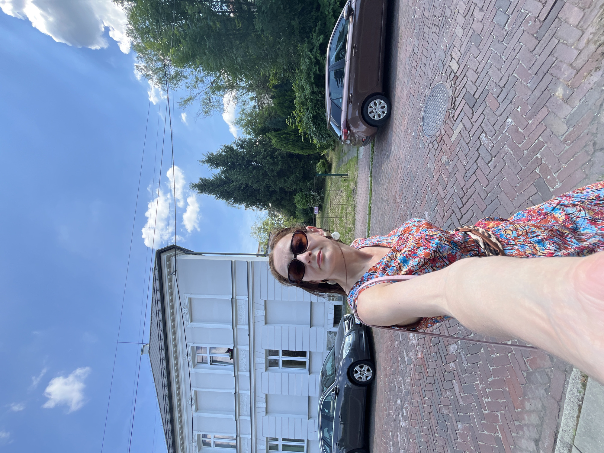The image is a selfie taken outdoors by a person wearing sunglasses and a colorful, patterned dress. The person is standing on a red brick sidewalk. Behind them, a white building with multiple windows and dark trim is visible. Two cars are parked on the street next to the sidewalk, one is dark grey and the other is a lighter brown. Trees and foliage line the background, with a bright blue sky with scattered white clouds above. The scene appears to be during the daytime with sunny weather.