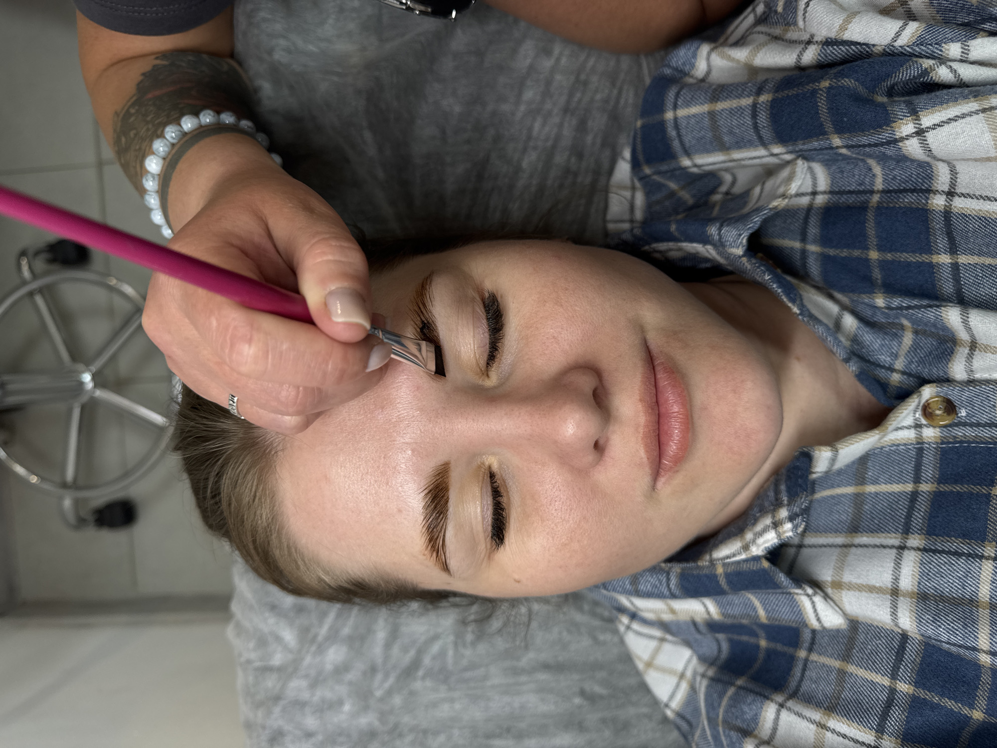 A woman lies on a gray-covered surface, wearing a blue and white plaid shirt. An individual with a tattoo on their arm, a white beaded bracelet, and a ring on their finger, is holding a pink-handled brush and a small silver tool to the woman's face, near her eyebrows. Her eyebrows are tinted darker, and her eyes are closed. A metal stool is partially visible in the background. There is no information to confirm the location in the city of Perm, Russia.