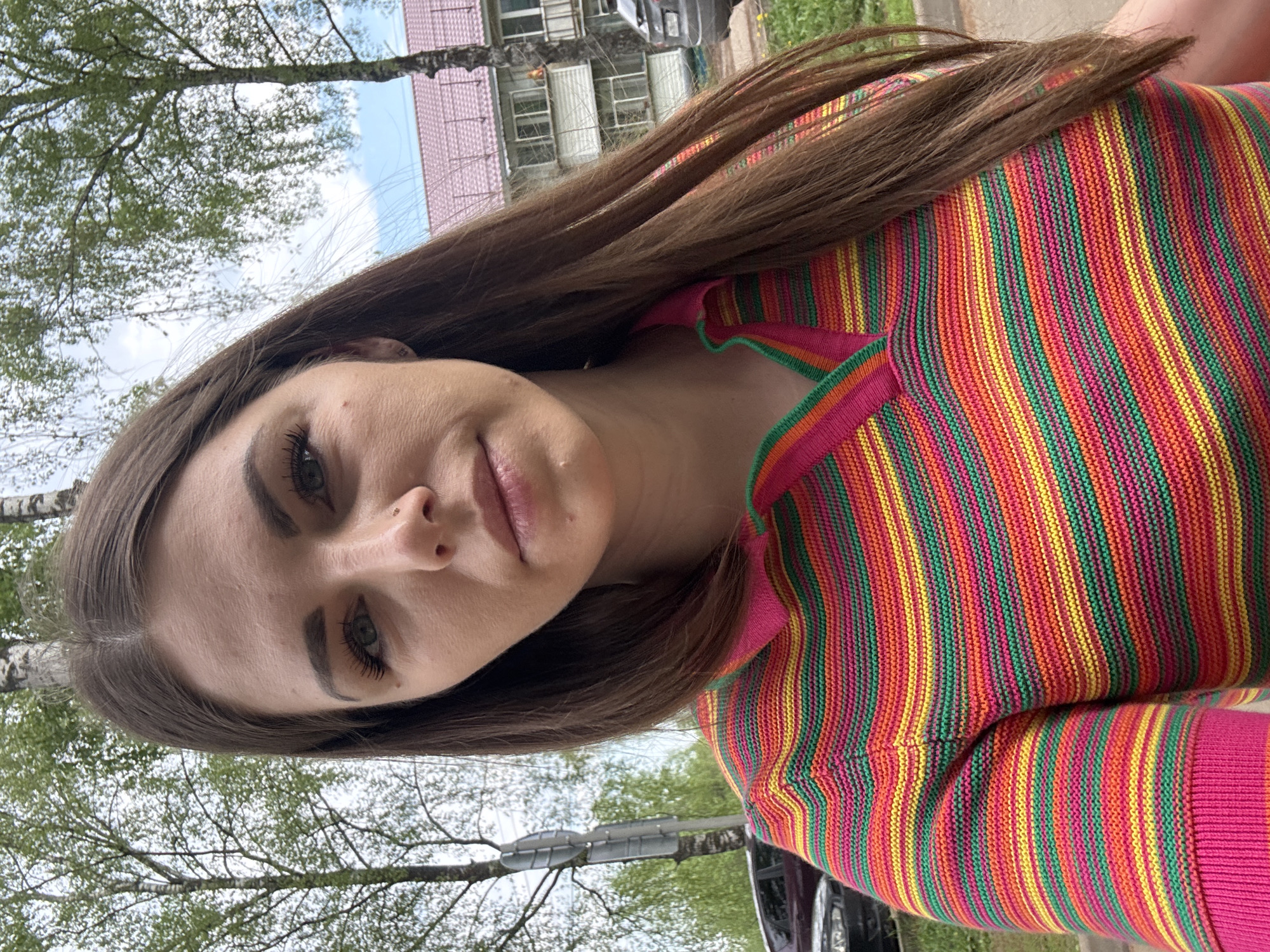 A woman with long brown hair is centered in a medium shot. She is wearing a brightly colored, vertically striped sweater in shades of pink, red, orange, green, and yellow. The background features trees with budding green leaves and a building with a pink roof and light-colored walls. A utility pole is also visible behind her. The photograph appears to have been taken outdoors during the daytime. Based on the surrounding background details, the location is likely within Раздолье, Russia.