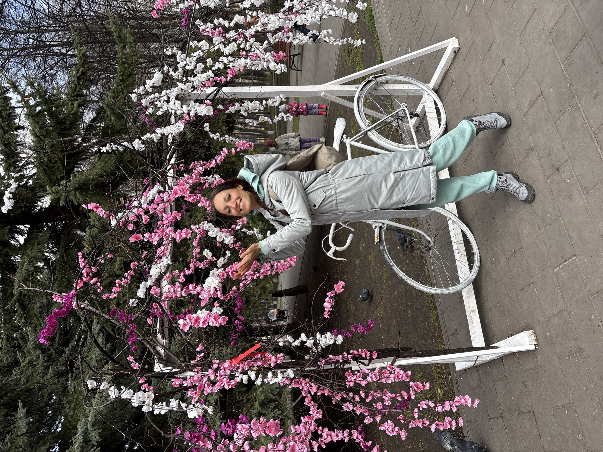 A woman smiles and poses next to a decorated white bicycle in a park in Ulyanovsk, Russia. The bicycle is adorned with artificial cherry blossom branches in shades of pink and white. The woman is wearing a grey coat, light blue tracksuit, and grey sneakers. She is reaching out towards the blossoms with her right hand. The background includes trees, other park decorations, and a brick pathway.