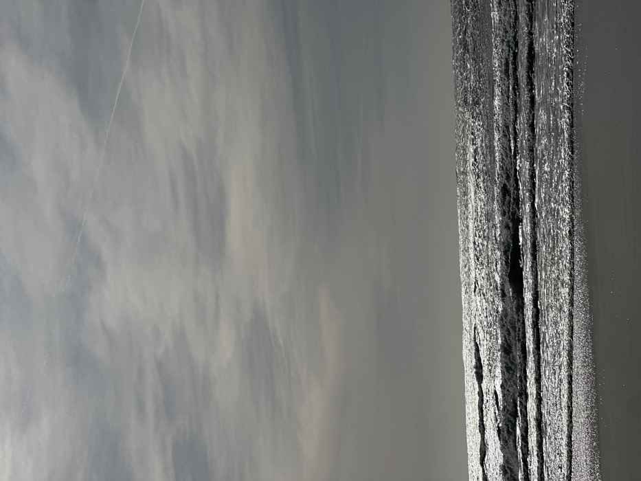 The image presents a coastal scene located in Gafanha da Nazaré, Portugal. The upper two-thirds of the frame consist of an overcast sky filled with light grey stratiform clouds. A single, distinct white linear aircraft contrail extends diagonally across the upper right quadrant of the sky. The lower third of the image displays the Atlantic Ocean, characterized by dark grey water with multiple parallel waves rolling towards the foreground. Intense sunlight reflection off the wave crests creates distinct shimmering horizontal bands of light across the water's surface. A narrow strip of dark, wet sand occupies the bottom edge of the frame, indicating the immediate shoreline. No individuals, animals, or man-made structures are present or discernible within the scene.