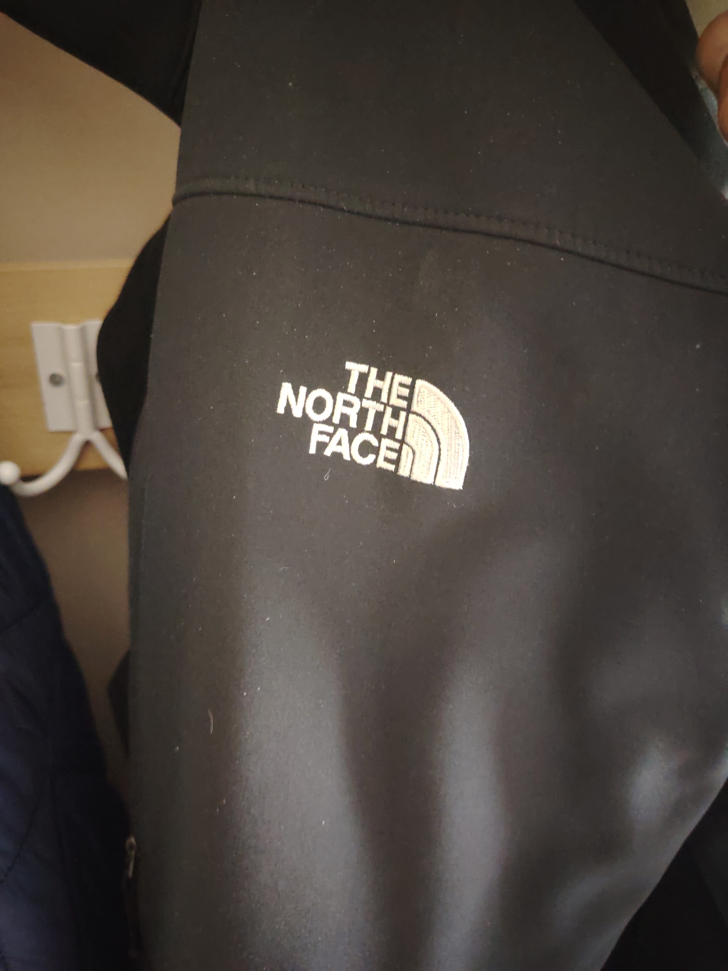 The image displays a black North Face jacket, identified by its white embroidered "THE NORTH FACE" logo and half-dome symbol, hanging within an interior space in Huntingdonshire, United Kingdom. The jacket's fabric surface exhibits minor particulate matter. A distinct horizontal seam with visible stitching traverses the garment. To the left of the jacket, a light-colored wooden surface is visible, upon which a white, two-pronged coat hook is mounted. A dark-colored garment hangs partially visible below the hook. The scene is illuminated by ambient lighting.