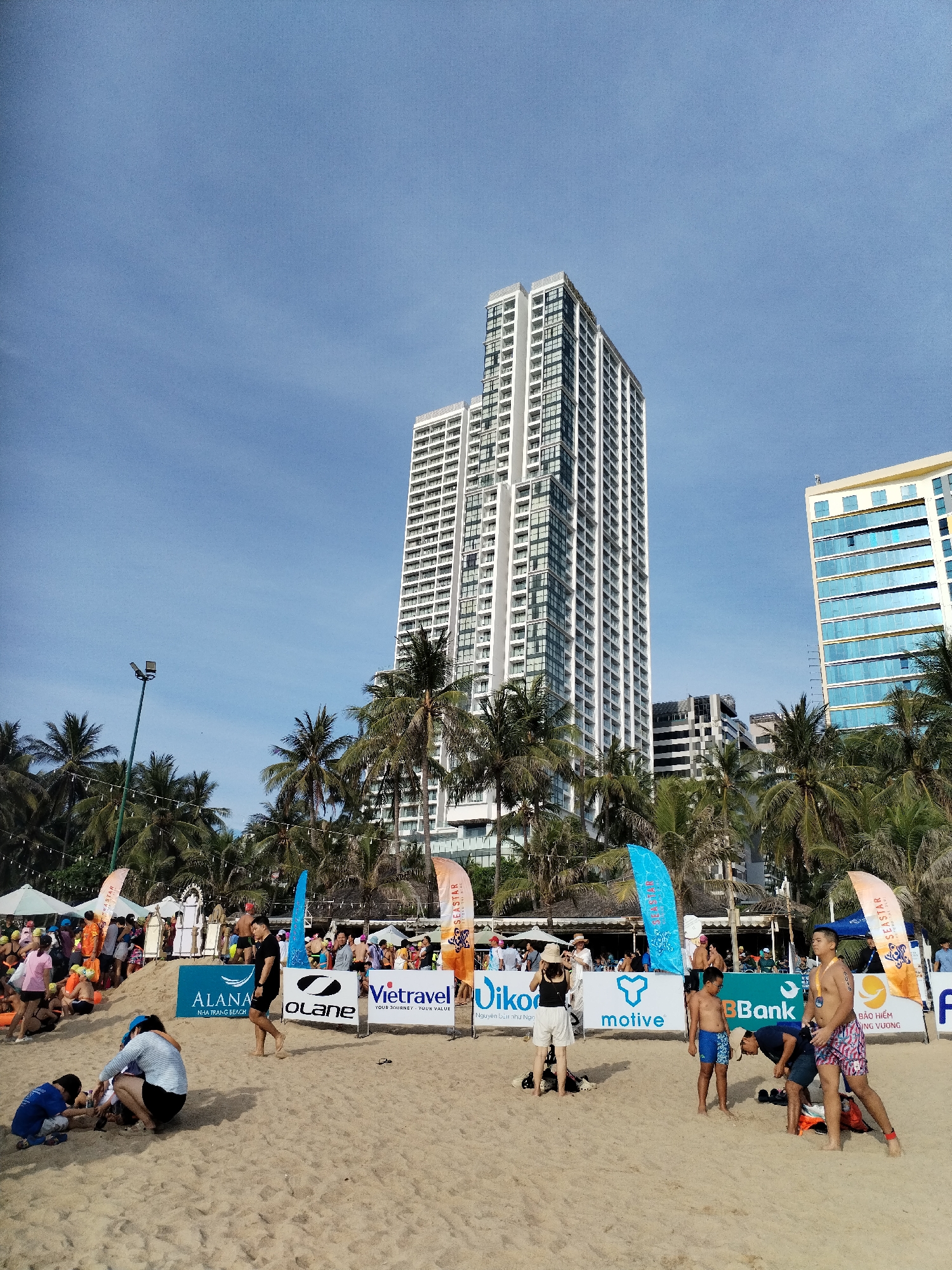 The image depicts a vibrant beach scene in Nha Trang, Vietnam, under a clear blue sky. In the background, two modern high-rise buildings dominate the skyline, one a tall, slender white skyscraper with numerous windows, and the other a wider, yellow-toned building with reflective blue windows. Coconut palm trees are scattered throughout the scene, framing the beach and the buildings.
The foreground is a sandy beach populated by a variety of people engaged in different activities. Several individuals are gathered near the water's edge, some appearing to be participating in an event, as indicated by colorful banners and flags with event branding. One prominent banner displays "Vietravel" with the tagline "YOUR JOURNEY. YOUR VALUE." Another banner features "OLANE" and "ALANA NHA TRANG BEACH."
On the sand, a group of children are playing, building with sand. Adults are seen observing, some standing and others sitting. A few individuals are dressed in swimwear. One man in blue patterned shorts is being attended to by another man who is bending down. A woman in a bikini is standing, looking to her right. Further back, a crowd of people are gathered under tents and along a beachfront promenade, suggesting a festival or organized event. The overall atmosphere appears to be lively and summery.