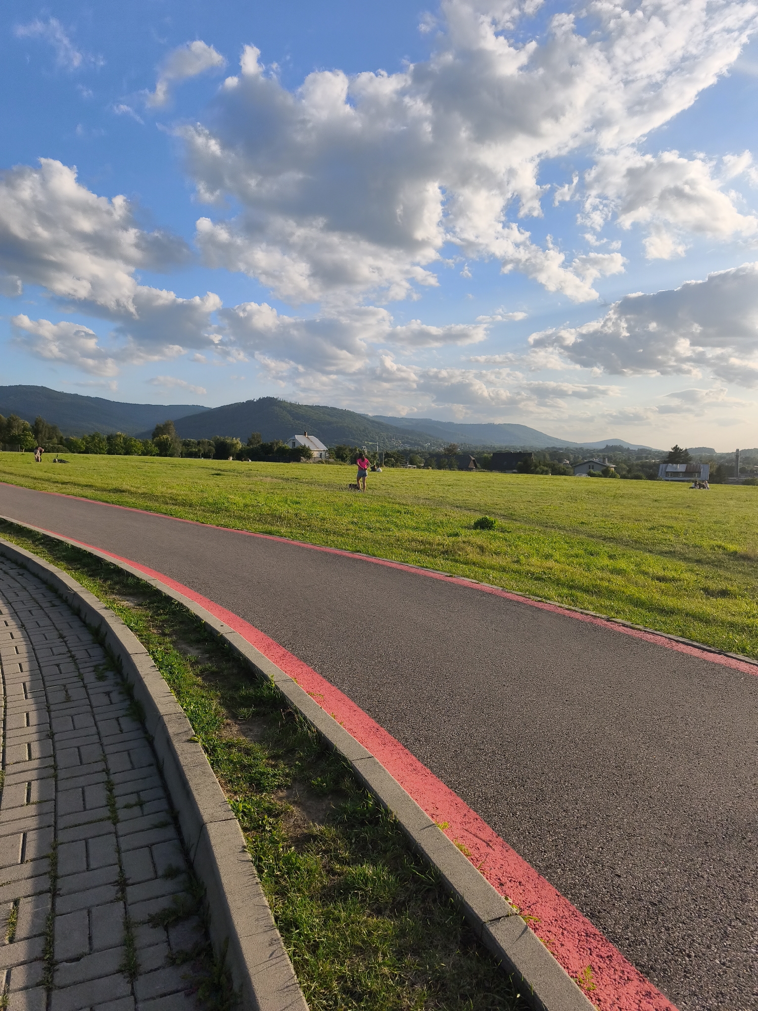 A paved pathway with a red border curves along the left edge of the frame, bordering a lush green grassy field. In the distance, rolling hills and mountains form the backdrop under a bright blue sky dotted with fluffy white clouds. Several people are scattered across the field, engaged in various activities. A person in a pink shirt stands near the right side of the frame, seemingly interacting with something on the ground, possibly a pet. Further to the left, another individual is visible, with objects that could be bicycles or other recreational equipment nearby. The scene appears to be a park or recreational area, with a tranquil and inviting atmosphere. The lighting suggests it's late afternoon or early evening, with the sun casting long shadows. The overall mood is peaceful and idyllic, showcasing a pleasant outdoor setting.