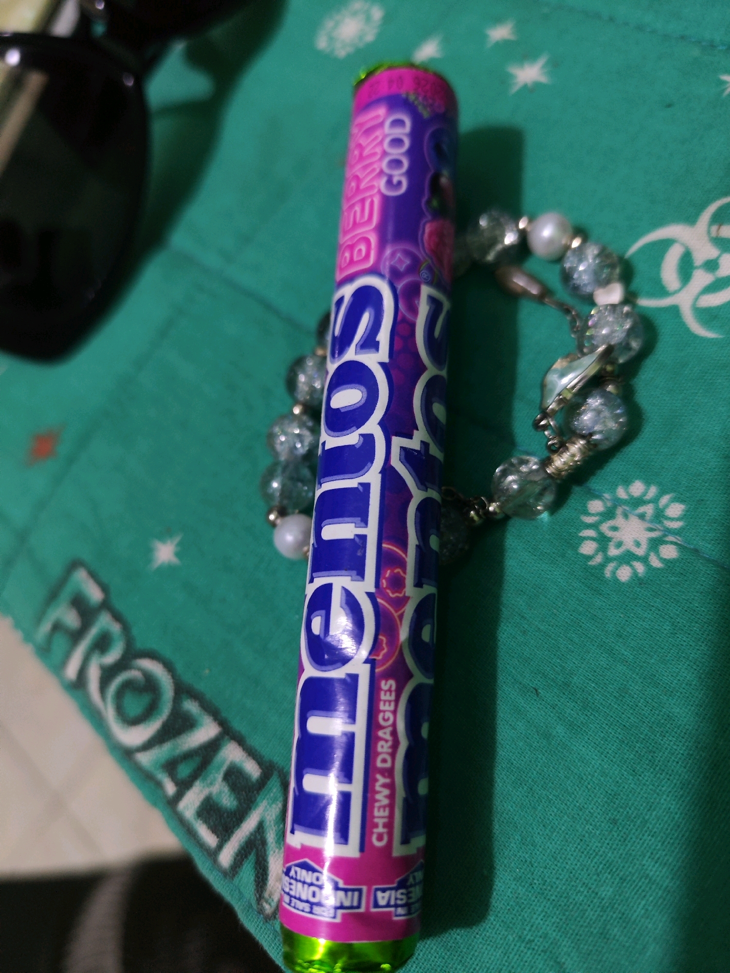 A purple and pink candy tube, labeled "BERRY GOOD" and "CHEWY DRAGEES," is positioned vertically in the center of the image. To the left of the candy tube, partially visible, is a black object, likely sunglasses. To the right of the candy tube is a beaded bracelet with light blue and white beads. The background is a teal-colored fabric with white snowflake-like patterns and the word "FROZEN" printed on it in white. The image appears to be taken indoors, as indicated by the lighting and shadow. There are no people visible in the image. The location is identified as Sroyo, Indonesia.