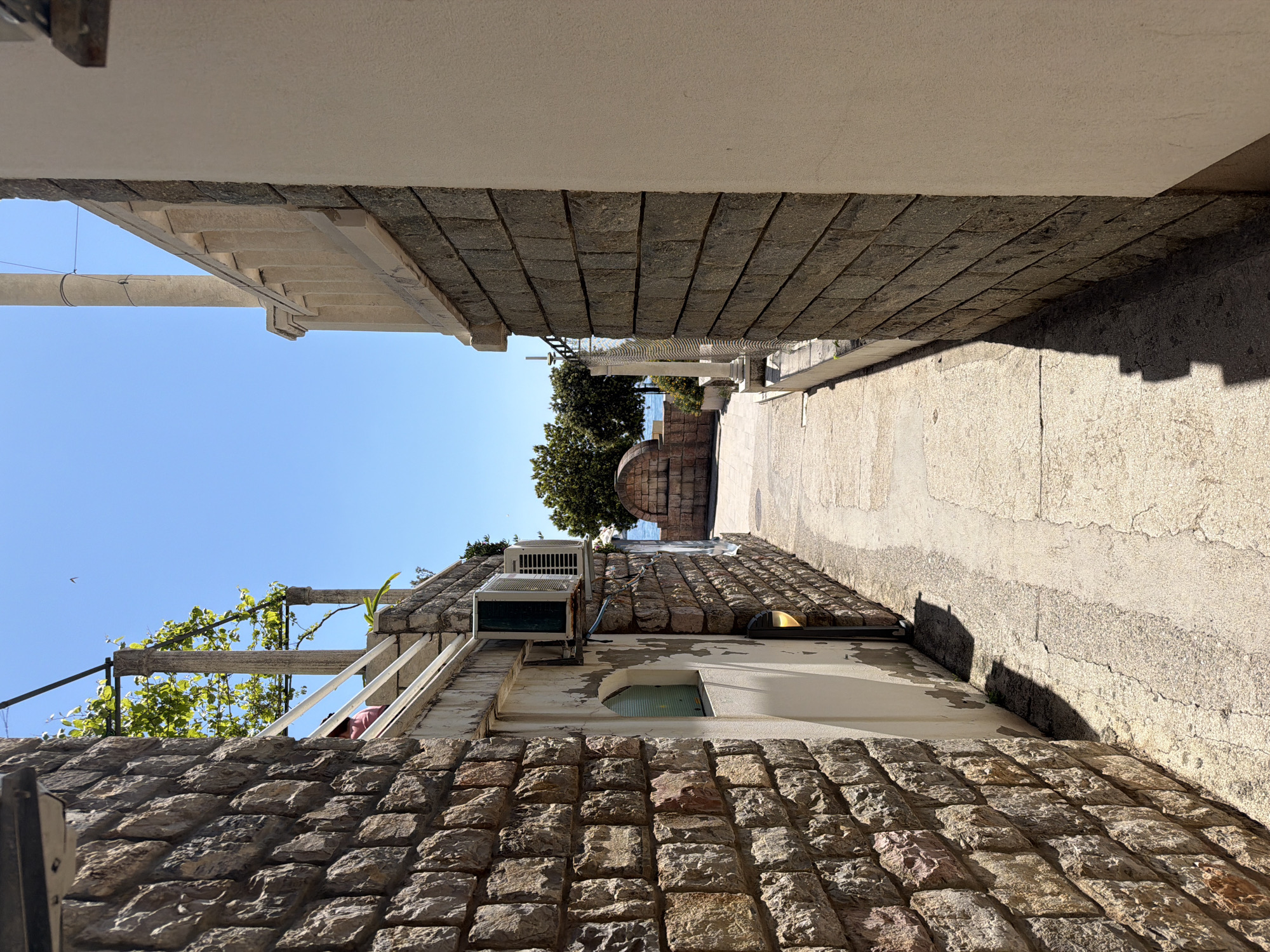 The image captures a narrow, sunlit alleyway between two buildings in what appears to be a coastal town, consistent with the provided location of Nerin, Montenegro. The perspective looks down the passage towards a distant view of the sea.
On the left, a building constructed with rough-hewn stone blocks rises from the ground. Above the stone section, the wall is lighter, possibly stuccoed, and features a small arched window. An air conditioning unit is mounted on this upper wall, and a wooden trellis adorned with green vines extends towards the clear blue sky.
The right side of the alley is also lined by a building with a stone base. A light-colored concrete or paved path runs alongside this structure. Strong sunlight casts sharp, elongated shadows from the building and an unseen overhead structure across the path, indicating a bright, sunny day, likely around midday or early afternoon. The ceiling above this path is a flat, light-colored surface, suggesting a covered area or an architectural overhang.
Further down the alley, beyond the immediate buildings, a glimpse of the calm, blue sea is visible under an equally clear blue sky. Some green trees and a section of a white, decorative balustrade or pillar can also be seen, contributing to the Mediterranean aesthetic of the scene.
No people or activities are visible in the image, presenting a quiet and still urban landscape. There is no visible text. The scene is dominated by the textures of stone masonry, the interplay of light and shadow, and the vibrant blue of the sky and sea, characteristic of a picturesque coastal setting.