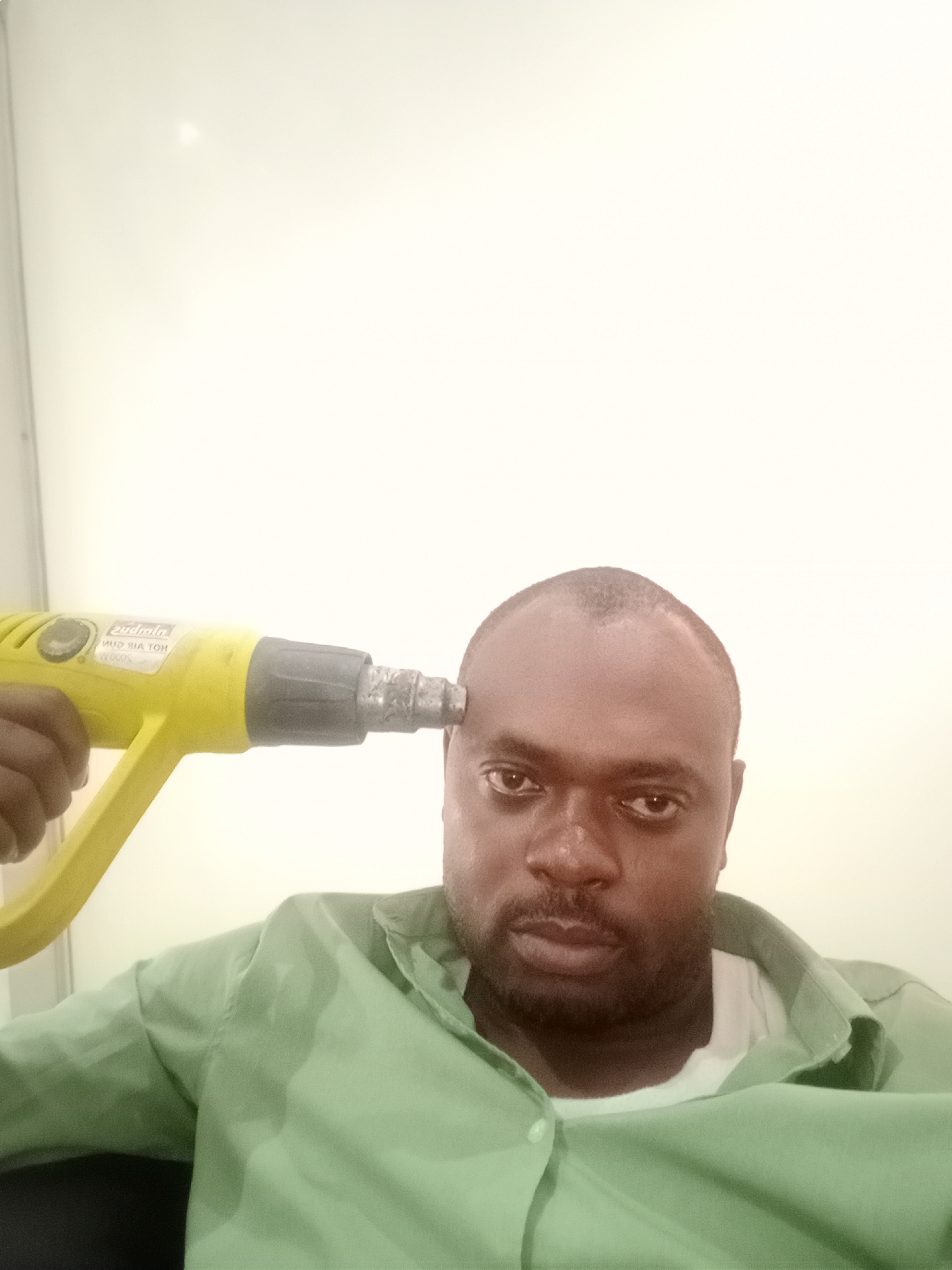 The image shows a man posing indoors, holding a yellow and grey heat gun to his right temple. The man has a dark complexion, short or shaved hair, and a beard. He wears a light green button-up shirt over a white t-shirt and looks directly at the camera with a neutral to slightly serious expression. His left hand is visible, gripping the heat gun's handle. The nozzle of the tool is pressed against his head, suggesting a humorous or symbolic gesture, possibly related to stress or intense thought. Visible, mirrored text on the heat gun includes "Sudalm," "HOT AIR GUN," and "2000W." The background is a plain, brightly lit, off-white wall, indicating an indoor setting with uniform lighting.