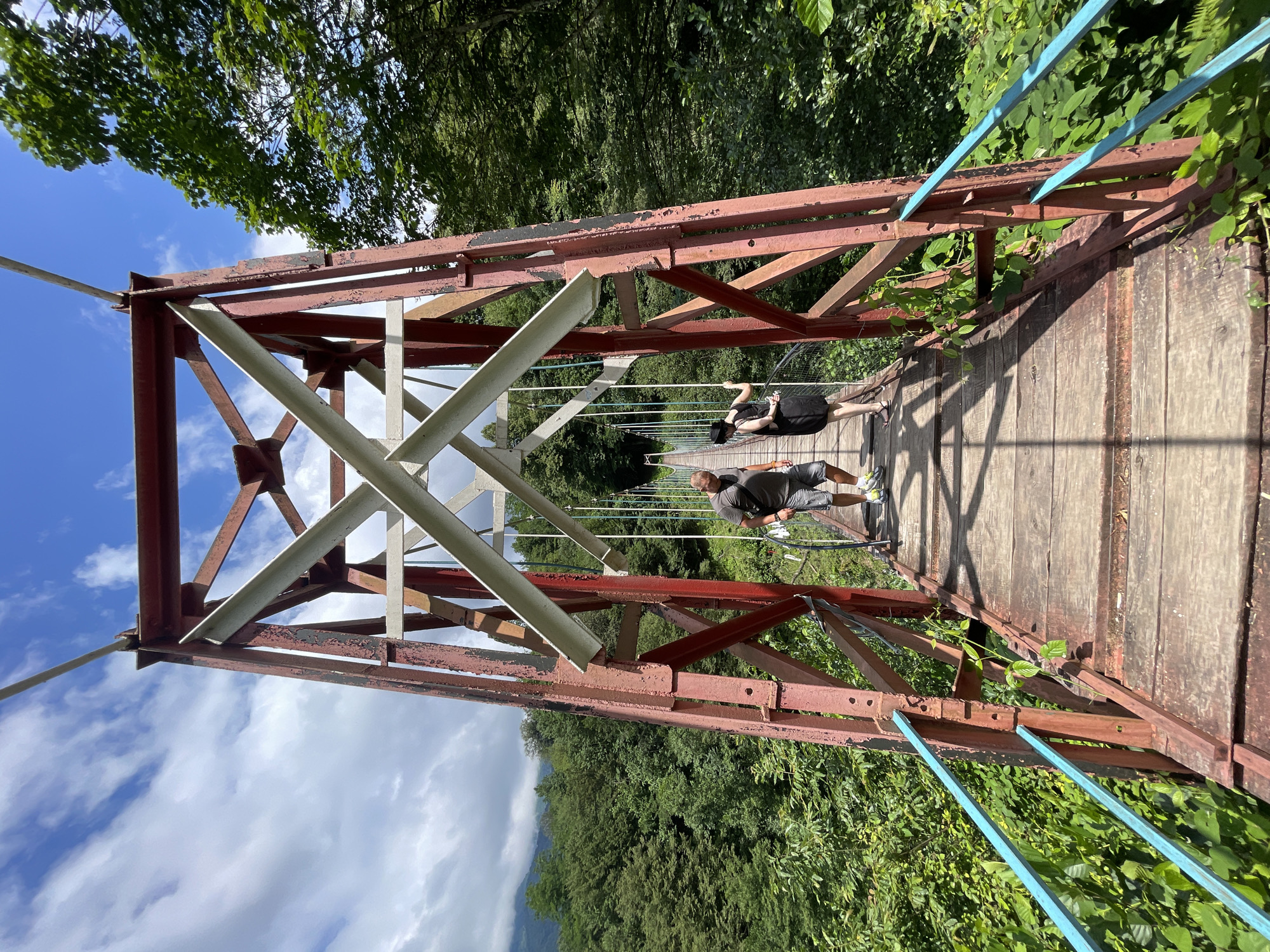 A medium shot captures a red suspension bridge in Tqwarchal, Abkhazia. The bridge, constructed of weathered wood and metal, spans a lush green gorge. Two individuals, a man and a woman, walk across the bridge from left to right. The man, wearing a grey t-shirt and shorts, is in front, while the woman, in a black dress and hat, follows closely behind. The surrounding environment is dense with trees and vegetation. The sky above is a mix of blue and white clouds.