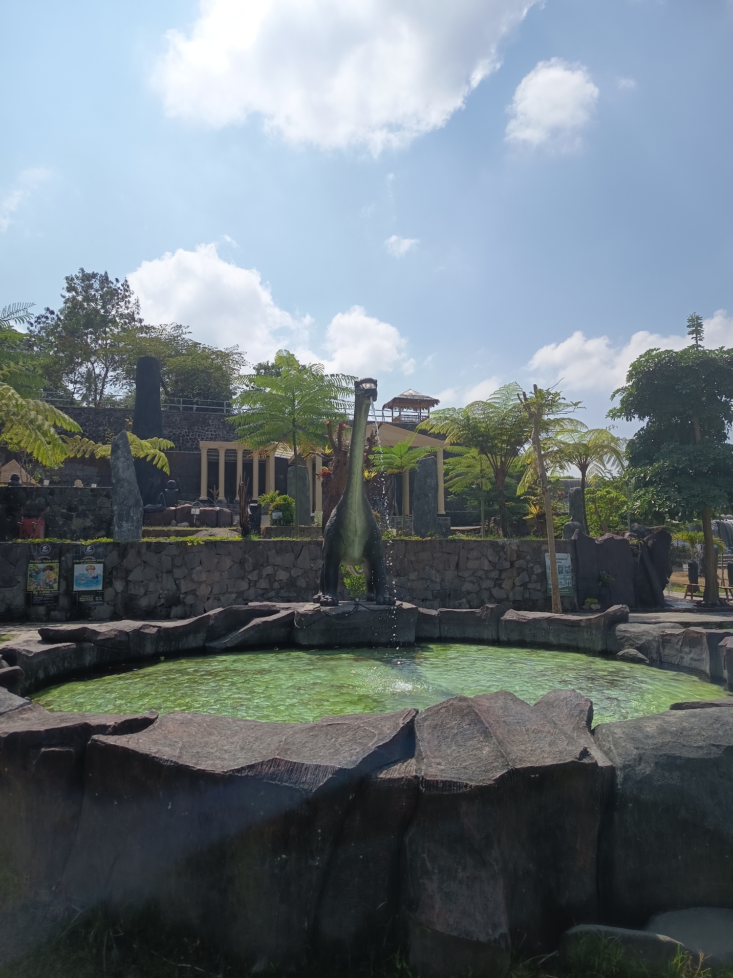This image, taken in the city of Sawangan, Indonesia, depicts an outdoor water feature with a dinosaur sculpture. A green-tinted pool of water is surrounded by large, dark, rock-like structures. In the center of the pool, a dinosaur sculpture with a long neck and tail is positioned, with water spraying from its mouth, creating a fountain effect. Behind the pool, a stone wall separates the foreground from a tiered landscape featuring more stone structures, columnar architecture, and lush greenery, including palm-like trees and ferns. A hut-like structure with a thatched roof is visible on an elevated level in the background. The sky above is blue with scattered white clouds. Signs, some with images of food and animals, are affixed to the stone wall. The overall scene suggests a park or attraction designed with a prehistoric theme.