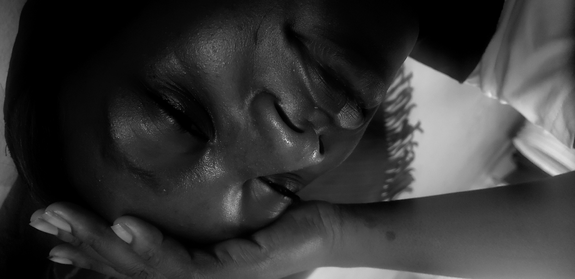 The image is a close-up, black and white photograph of a person's face resting on a hand. The person's eyes are closed, and their facial features are soft. The skin appears smooth, possibly moist. The hand supporting the face has well-manicured nails. Part of an arm extends into the frame. The background is blurry and out of focus, showing hints of a textured surface and a light-colored object, possibly fabric. There is no indication or evidence in the image that this scene takes place in Narayi, Nigeria, or any specific location.