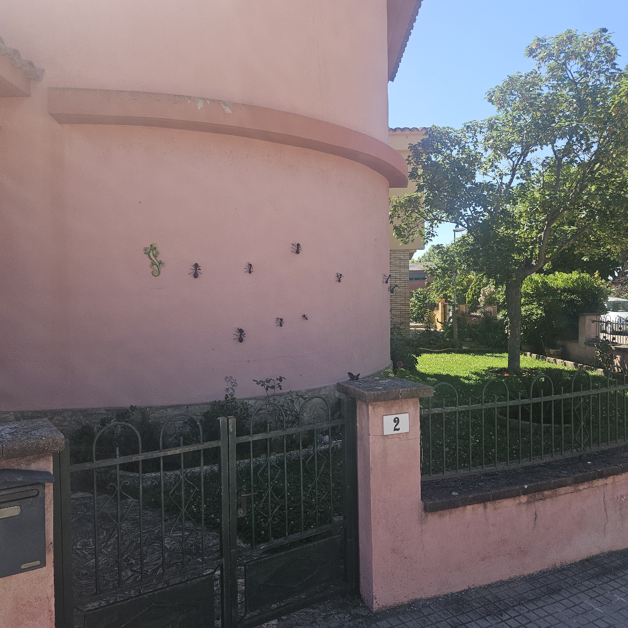 The image shows the exterior of a building in La Jonquera, Spain. The building has a pink facade adorned with decorative metal ants and a lizard. A wrought iron gate and fence surround a garden with green lawn and various plants and trees. A house number "2" is affixed to a pillar near the gate. The sky is blue and clear. A mailbox is attached to the left pillar.