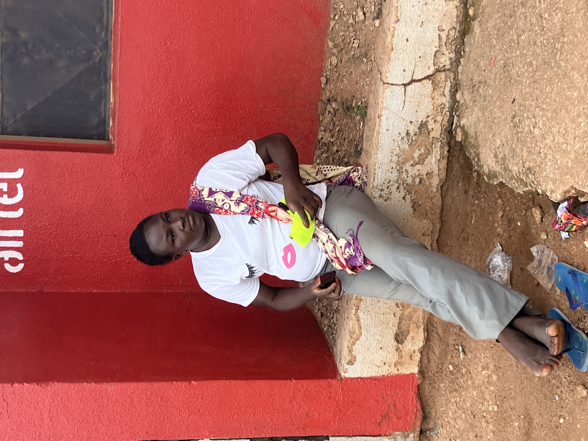 In Gura Top, Nigeria, a person with dark skin sits on a concrete ledge against a bright red building. The person is wearing a white t-shirt with a graphic of eyelashes and lips, gray pants, and a colorful patterned scarf draped over their shoulders. They are holding a neon yellow cup in their right hand and a small dark object in their left. Their feet, adorned with a toe ring, are bare and resting on a pair of blue flip-flops. The ground is made of dirt and small stones, with some plastic debris visible. A portion of a window is visible on the red building, and white text that appears to be "Airtel" is also present on the wall.
