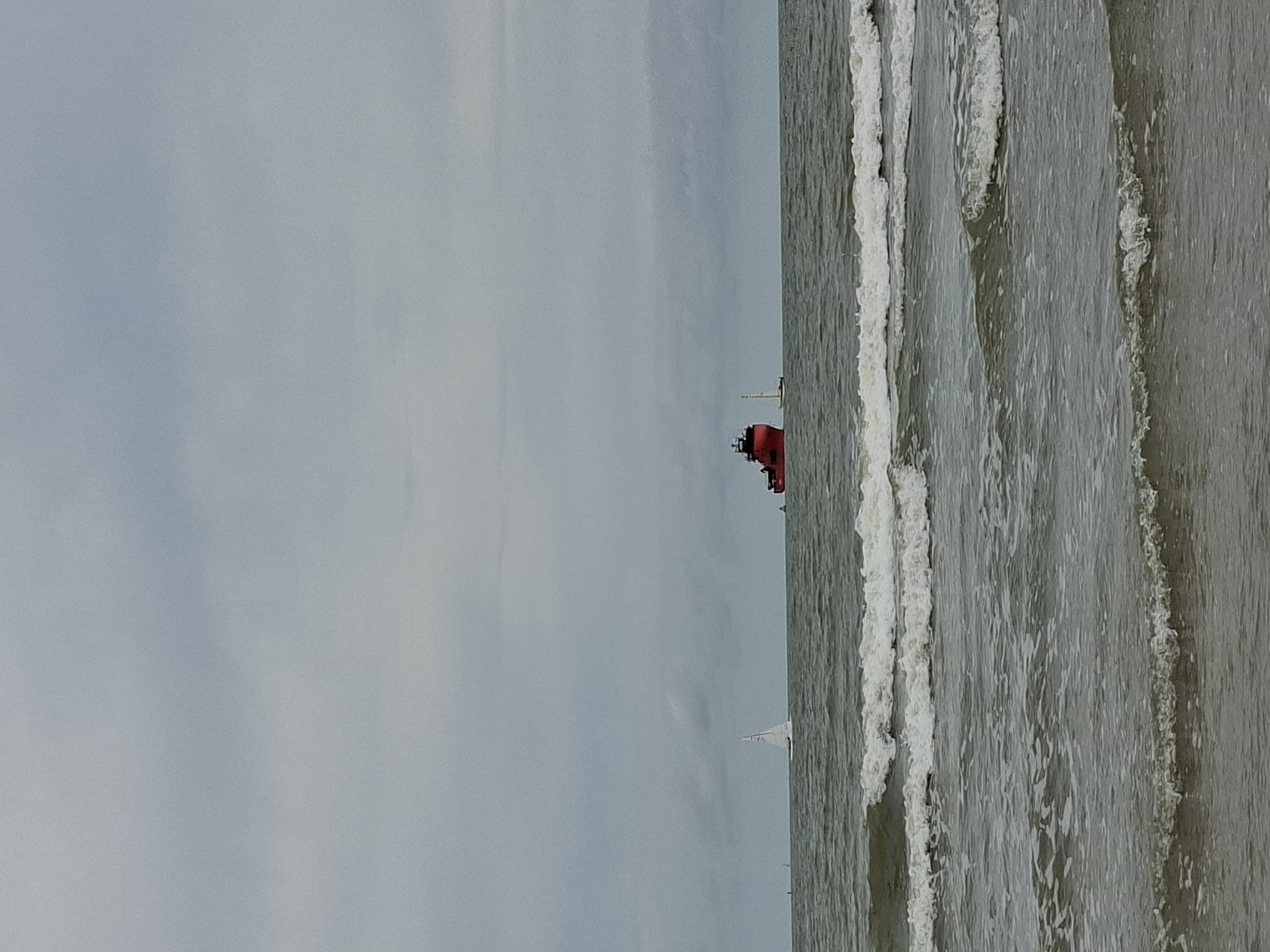 This image depicts the North Sea off the coast of Ostend, Belgium, under an overcast sky. The scene is dominated by choppy, grey water with white-capped waves breaking near the shore. In the distance, a large red cargo ship is visible. To the left of the cargo ship, a white sailboat is sailing. Further to the left, another smaller sailboat can be seen. The horizon line is indistinct due to the hazy weather. The foreground is occupied by the beach and the incoming waves. There are no people clearly visible in the image. The overall atmosphere is one of a typically cloudy day at sea.