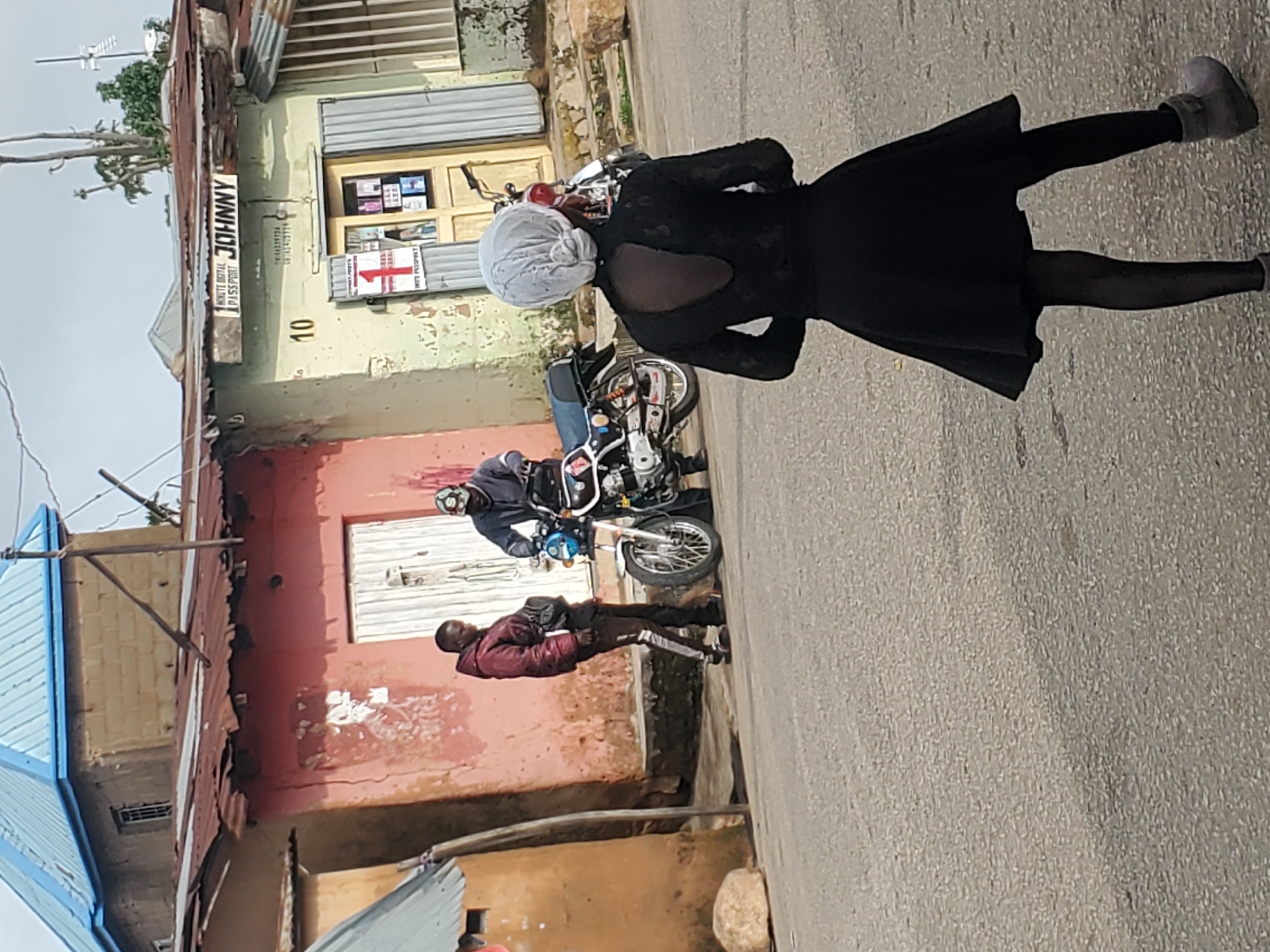 In the city of Jos, Nigeria, on an overcast day, a street scene unfolds. A woman wearing a black dress, black tights, and grey boots stands facing away from the camera, her head covered by a white headscarf tied in a bun. To her left, two men are near a motorcycle. One man, wearing a maroon jacket and black pants with a white stripe, has a black fanny pack slung across his chest. The other man, wearing a dark jacket and a baseball cap, is seated on the motorcycle, which is white and black with blue accents. Behind the men is a building with a corrugated metal roof, a light green painted facade, and a sign that reads "JOHNNY" above a smaller sign reading "MINUTE DIGITAL PASSPORT." The building also has a numbered address "10" and a closed wooden door with frosted glass panels displaying framed items. To the left of this building is another structure with a faded pink facade and a corrugated metal roof. A stone sits on the roadside in the foreground. The ground is paved with asphalt.