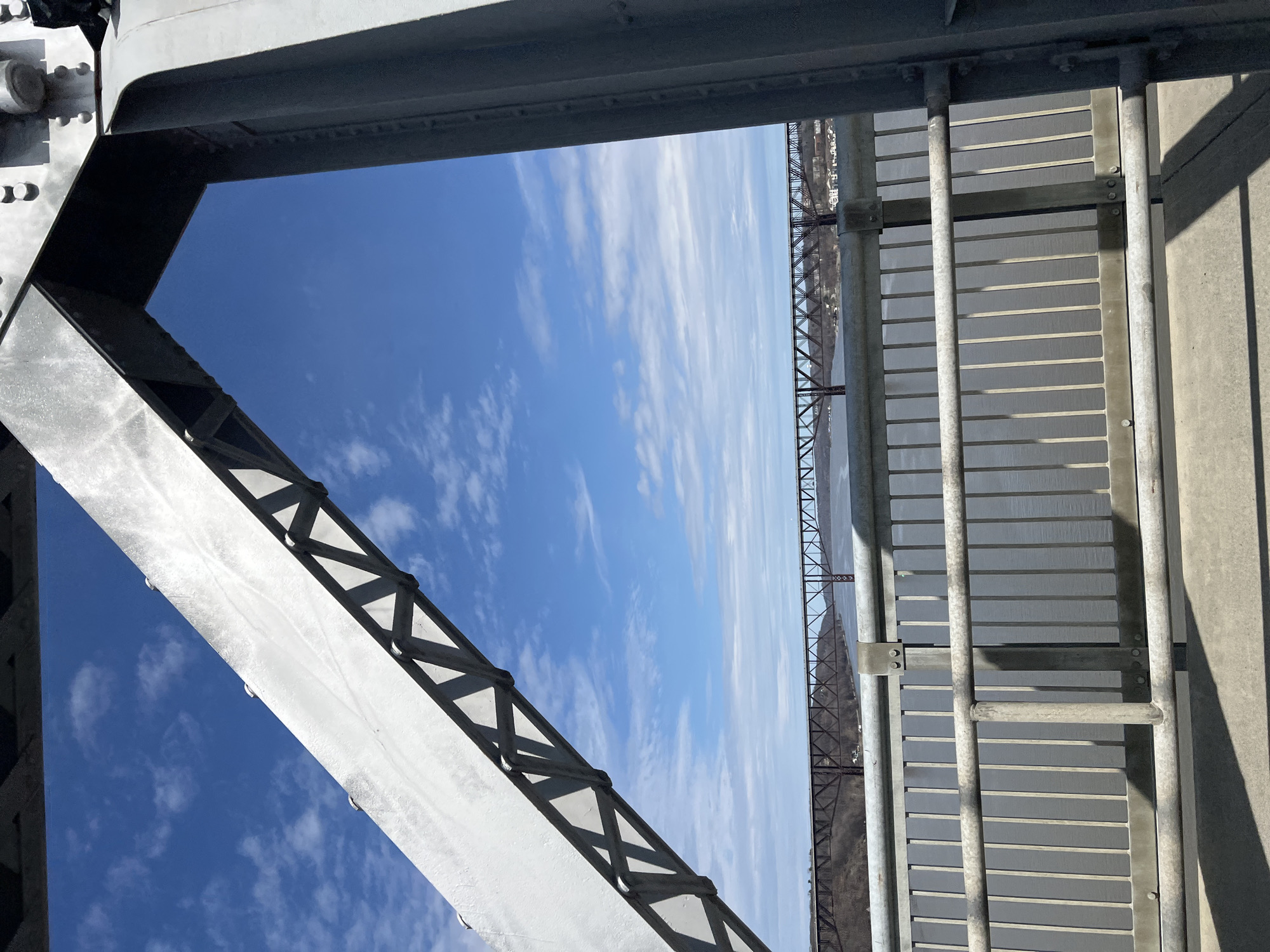 The image depicts a view through the structural elements of the Walkway Over the Hudson in Poughkeepsie, New York. The scene includes sections of the bridge's steel framework, walkway barriers, and a distant view of the Hudson River and its surrounding landscape. The sky is blue with scattered clouds. The bridge's structure is grey and metallic, with various connecting joints and supports visible. The river appears as a light blue band in the distance.