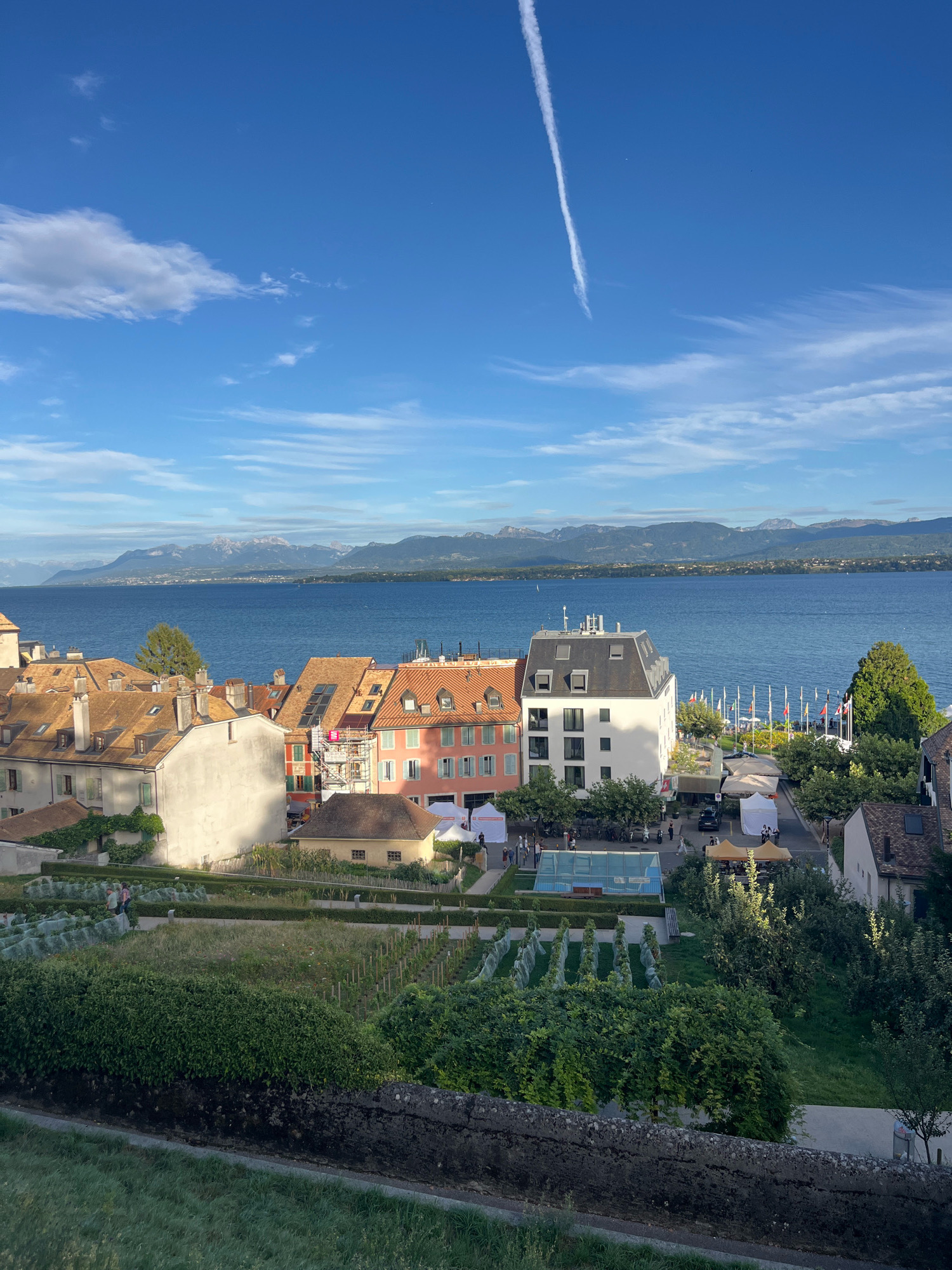 The image depicts a scenic view of Nyon, Switzerland, overlooking Lake Geneva and the distant Alps. In the foreground, a neatly manicured garden with rows of plants and trees is visible. Beyond the garden, a cluster of European-style buildings with terracotta roofs line a street. A modern white building with a dark sloped roof stands out among them. Along the lakefront, numerous flags are displayed, suggesting a public space or event. The lake itself is calm, reflecting the clear blue sky. A faint contrail from an aircraft streaks across the sky. Several people are observed walking around the garden and street, with some small tents or stalls set up, possibly for a market or event. The overall impression is one of a peaceful, picturesque town on a sunny day.