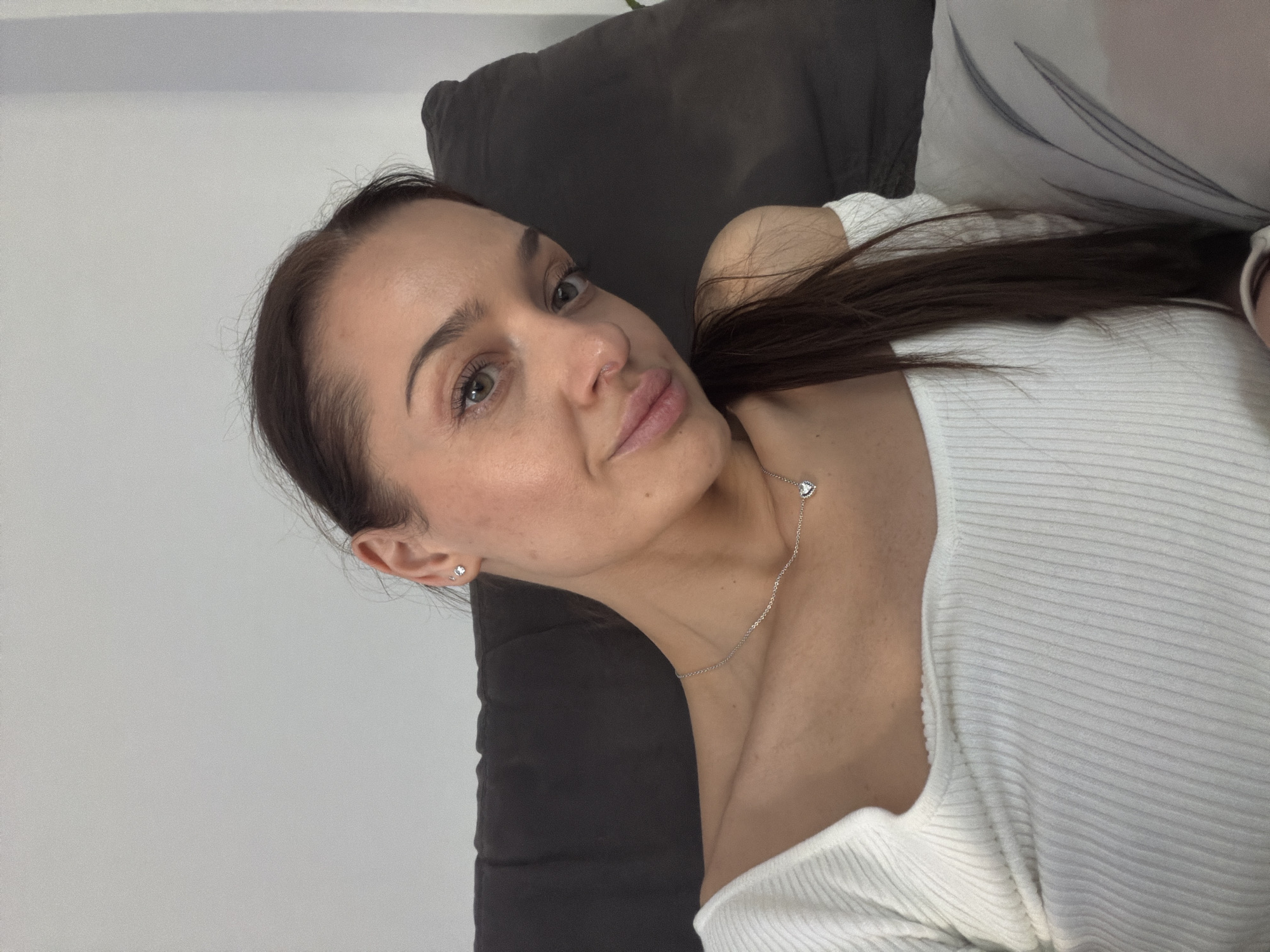 A woman with brown hair and light skin is lying on a dark gray couch. She is wearing a white, ribbed, low-cut shirt and a delicate silver necklace with a heart-shaped pendant. She is also wearing small stud earrings. The background is a plain white wall. The scene appears to be indoors. There is no indication that the image was taken in Varna, Bulgaria.