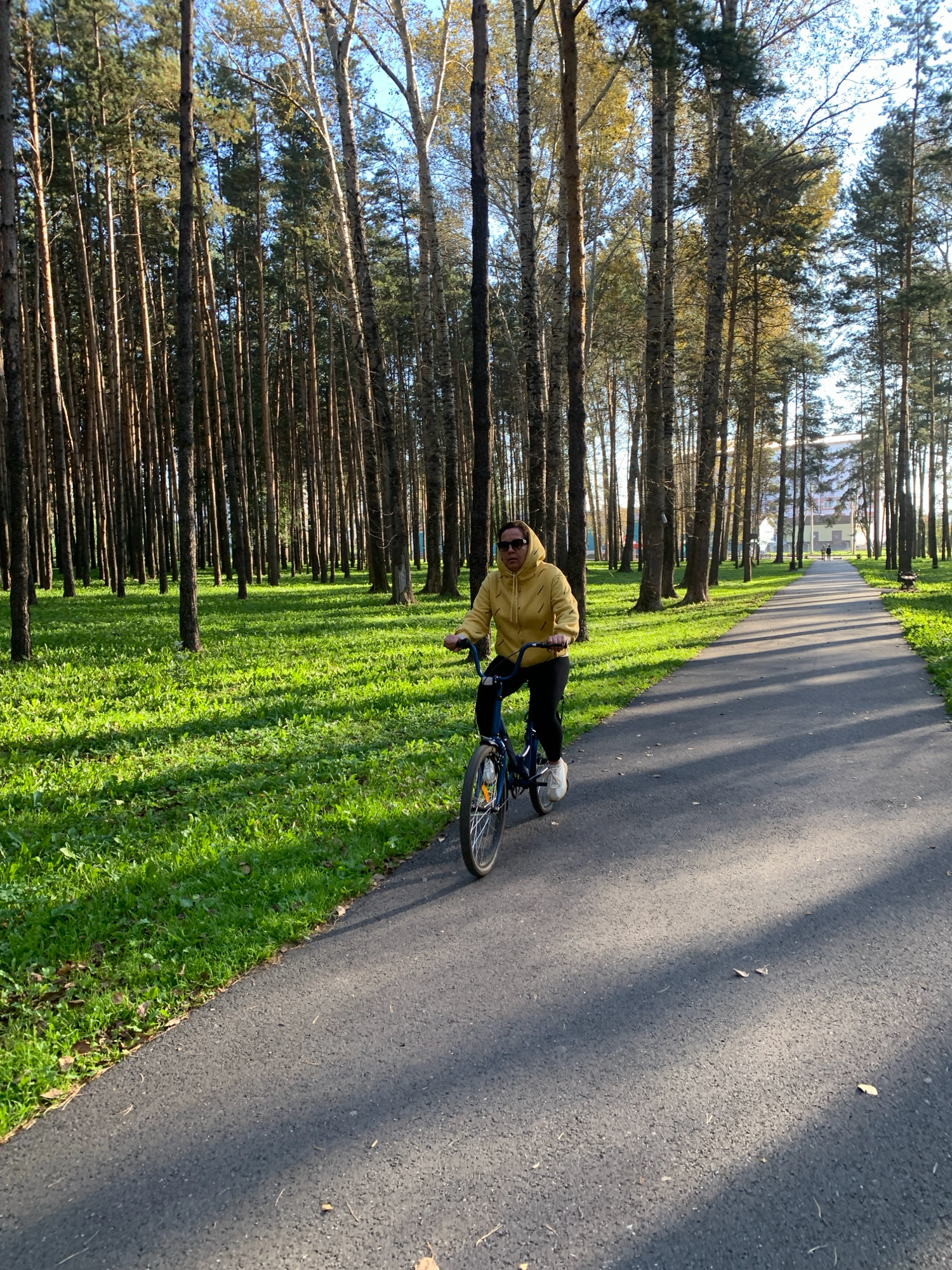 An adult female wearing sunglasses and a yellow hooded jacket rides a blue bicycle along an asphalt path. The path is bordered by a grassy area on the left and tall, slender trees on the right. The sun casts shadows from the trees across the path and the grass. In the distance, there is a building and another person walking. The scene appears to be a park in the city of Prokopyevsk, Russia.