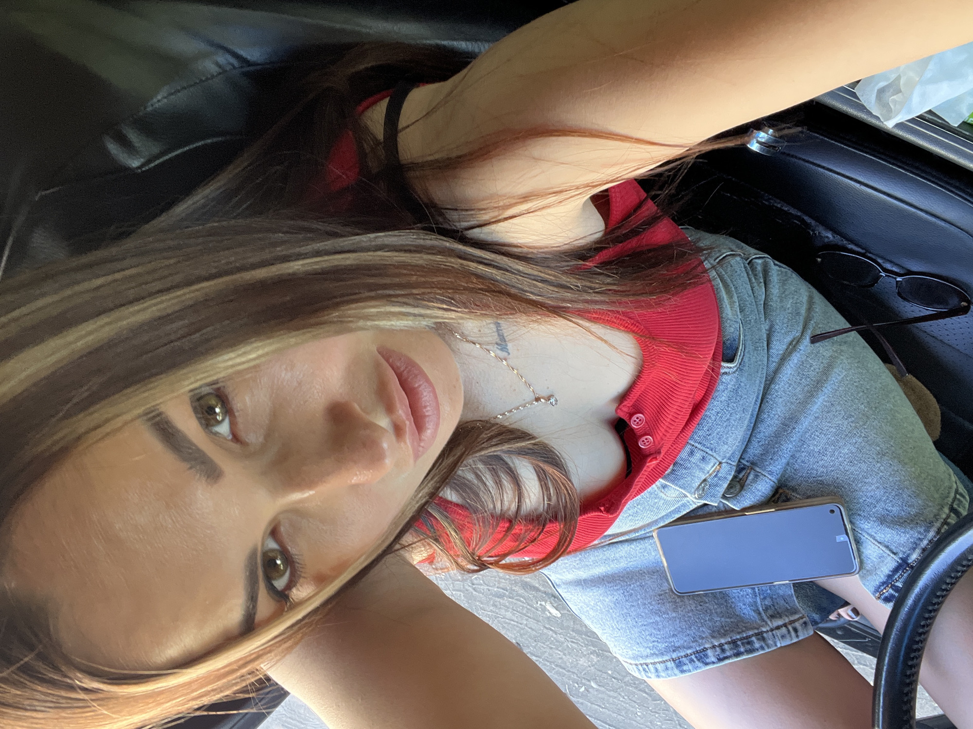 A woman with long, layered brown hair with blonde highlights is sitting in a car, looking directly at the camera. She is wearing a red tank top and a denim skirt. A smartphone is resting on her lap, and a pair of sunglasses is on the car seat next to her. The background appears to be the interior of the car, with black leather seats visible. The lighting suggests it is daytime, possibly sunny, as there is a strong light source and shadows.