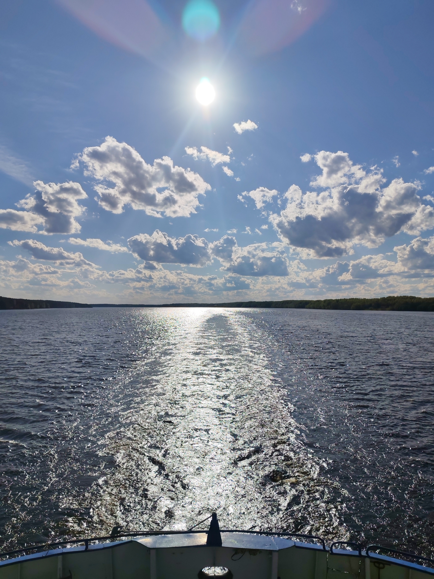 The image depicts a view from a boat on a body of water, possibly the Volga River near the city of Myshkin, Russia, given the context of the image. The sun is high in the sky, partially obscured by light clouds, creating a glare on the water's surface. The reflection of the sun forms a bright, elongated path on the water leading to the horizon. The shoreline is visible in the distance with a line of trees. The bow of the boat is visible at the bottom of the frame, with the metal railing and part of the deck in view. No people are visible in the shot.